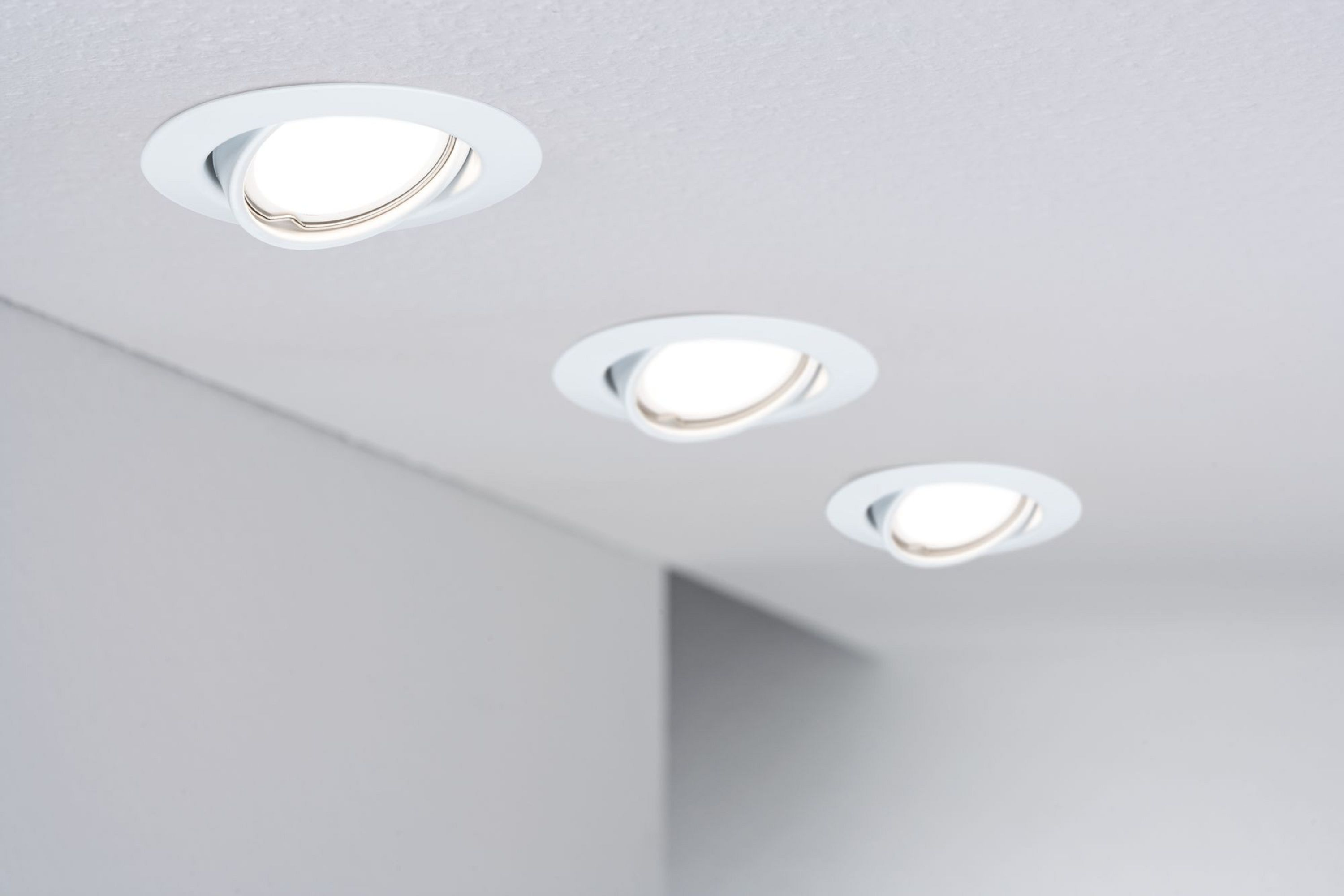 Drei weiße Einbau-Deckenleuchten sind beleuchtet, abgewinkelt und vor einer weißen Decke angeordnet. Die Wand ist hellgrau.