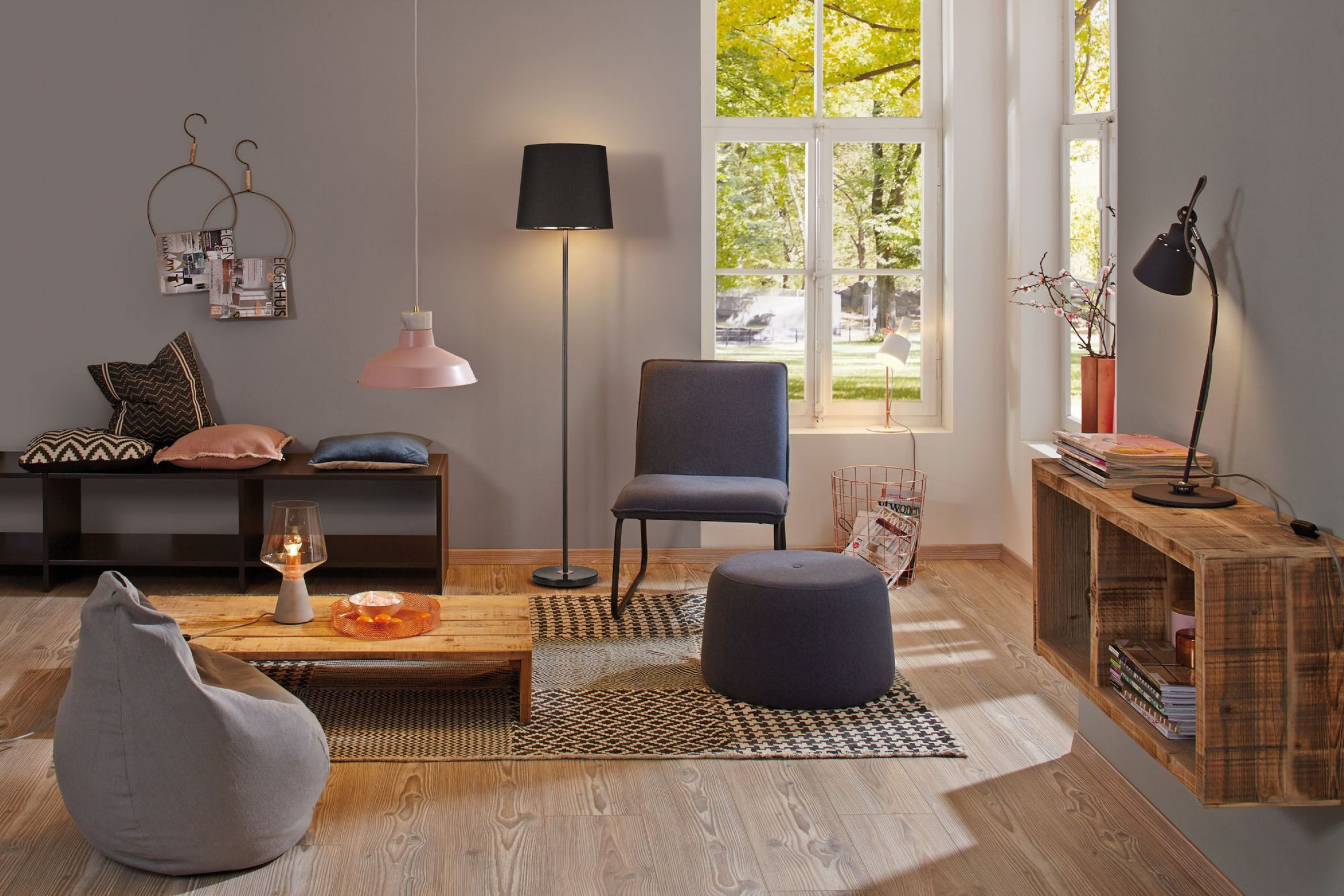 Przytulny salon z krzesłem, pufą, lampami, ławką z poduszkami, czasopismami, drewnianym stołem i oknem z drzewami.