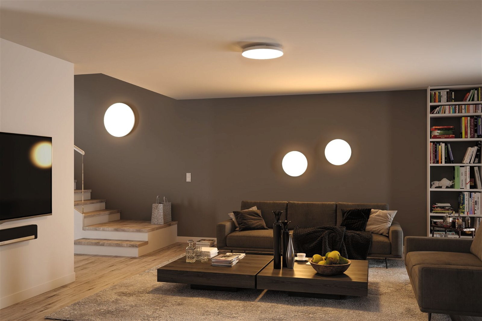 Wohnzimmer mit Sofa, Couchtisch, Lichtern und Bücherregal. Treppen sind im Hintergrund zu sehen, links ein Fernseher.