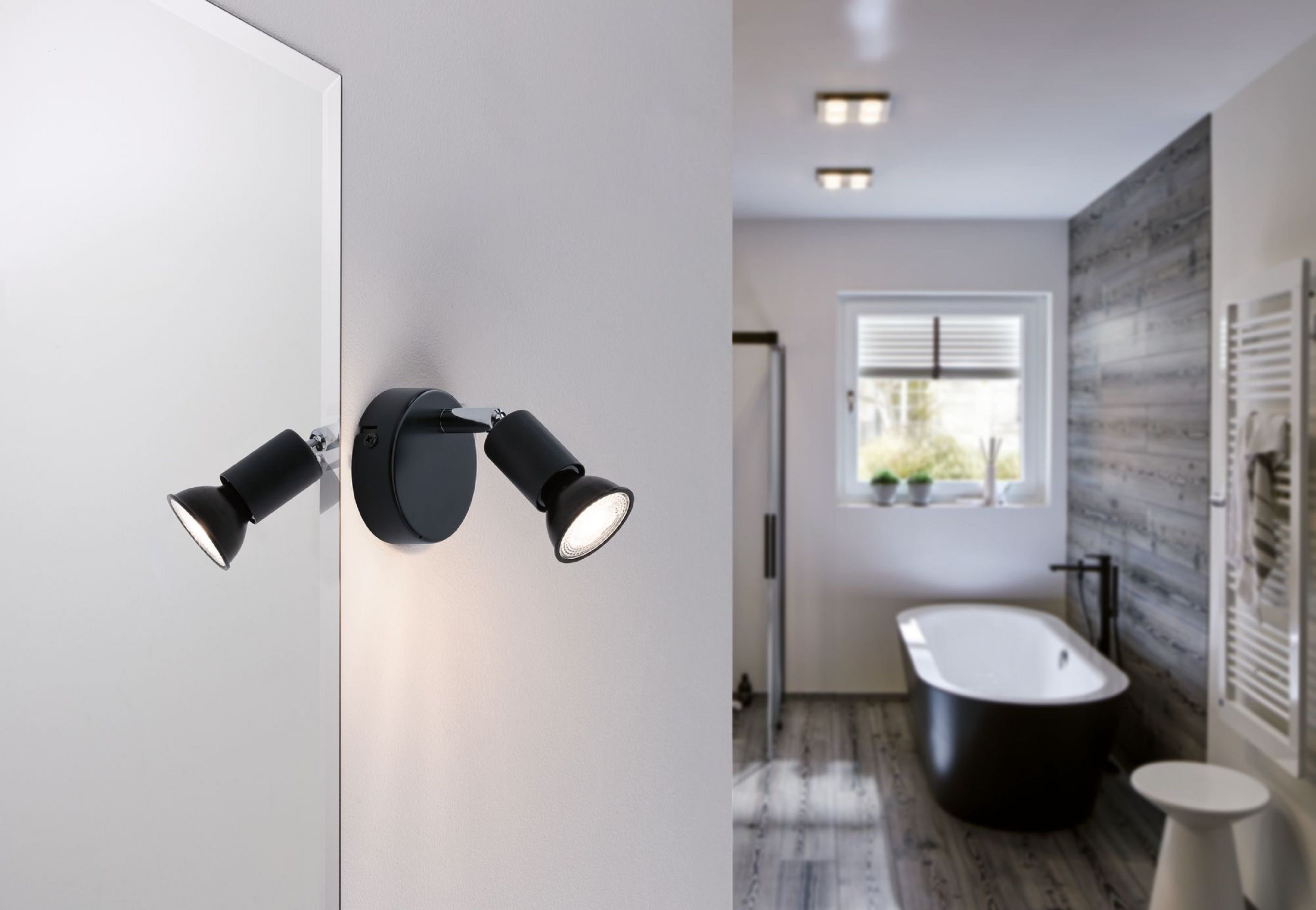 Schwarze Strahler an einer weißen Wand beleuchten ein Badezimmer. Im Hintergrund befinden sich eine Wanne und ein Fenster.