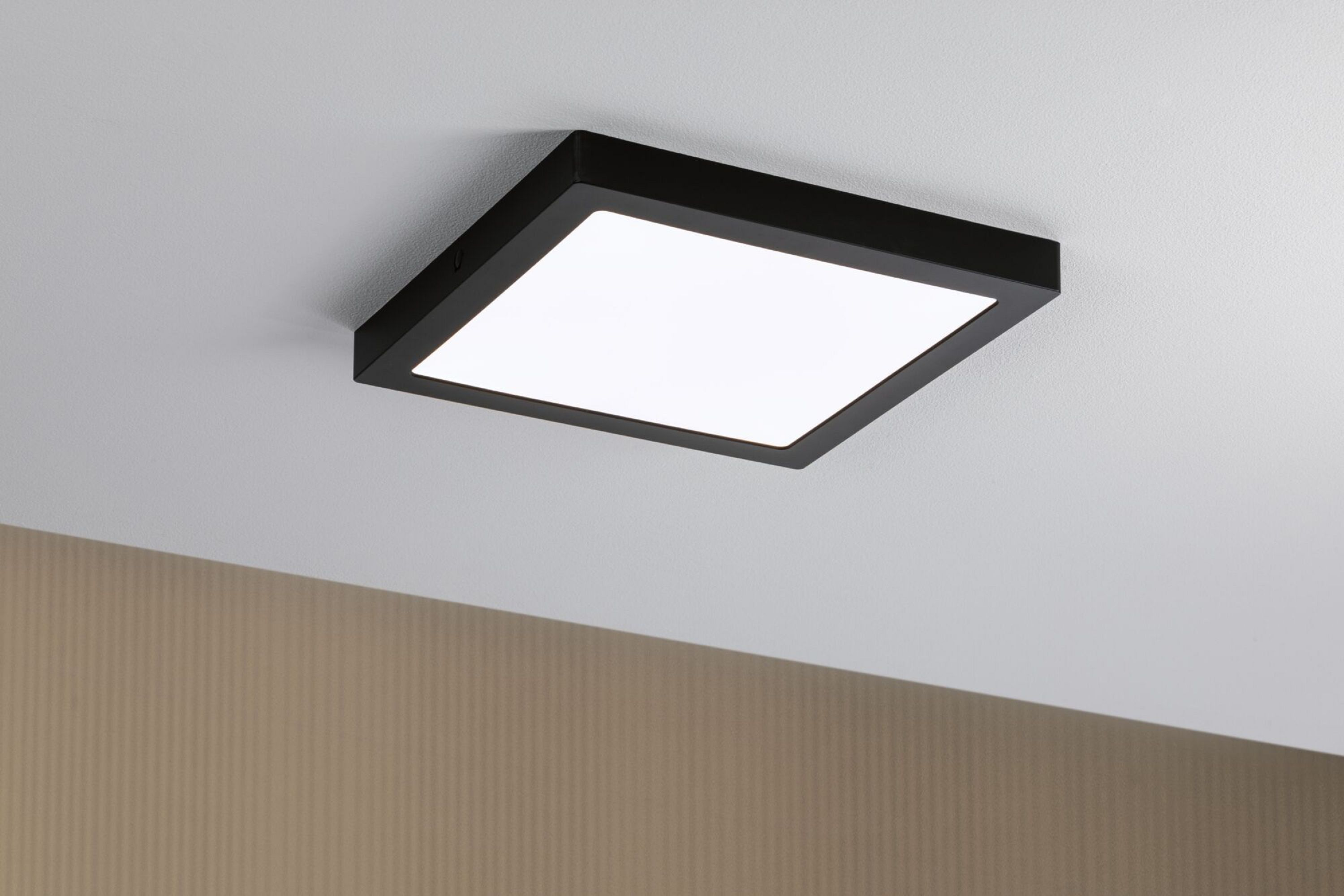 Una plafoniera quadrata nera con una luce bianca brillante, montata su un soffitto bianco. Un muro beige è nell'angolo.