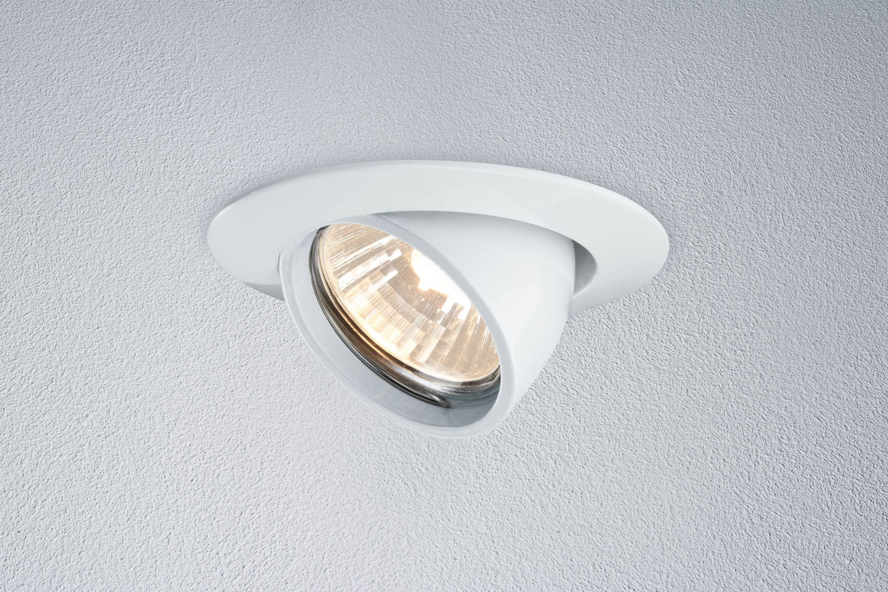 Ein weißer Einbaustrahler ist abgewinkelt. Er ist in einer weißen Decke eingesetzt. Das Licht ist an.