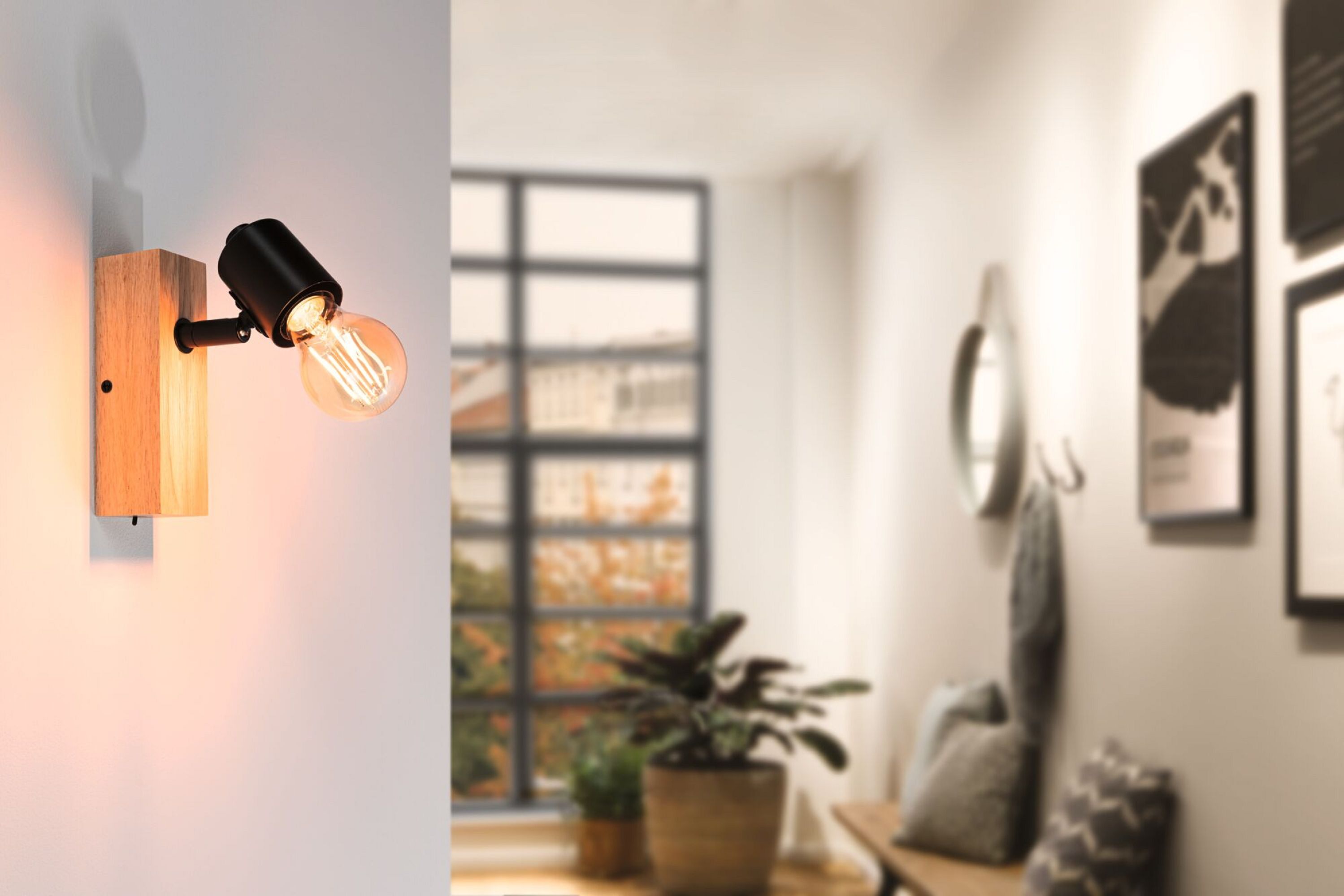 Lampa ścienna z żarówką Edisona świeci na drewnianej podstawie. Korytarz z roślinami, lustrem i dziełami sztuki w tle.