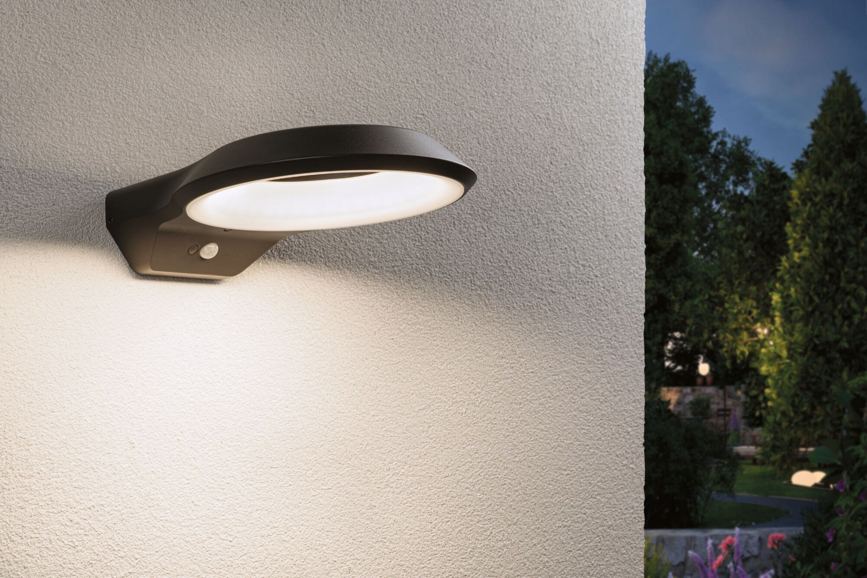 Czarna, owalna lampa na ścianie oświetla teksturowaną ścianę. W tle widoczny wieczorny ogród.