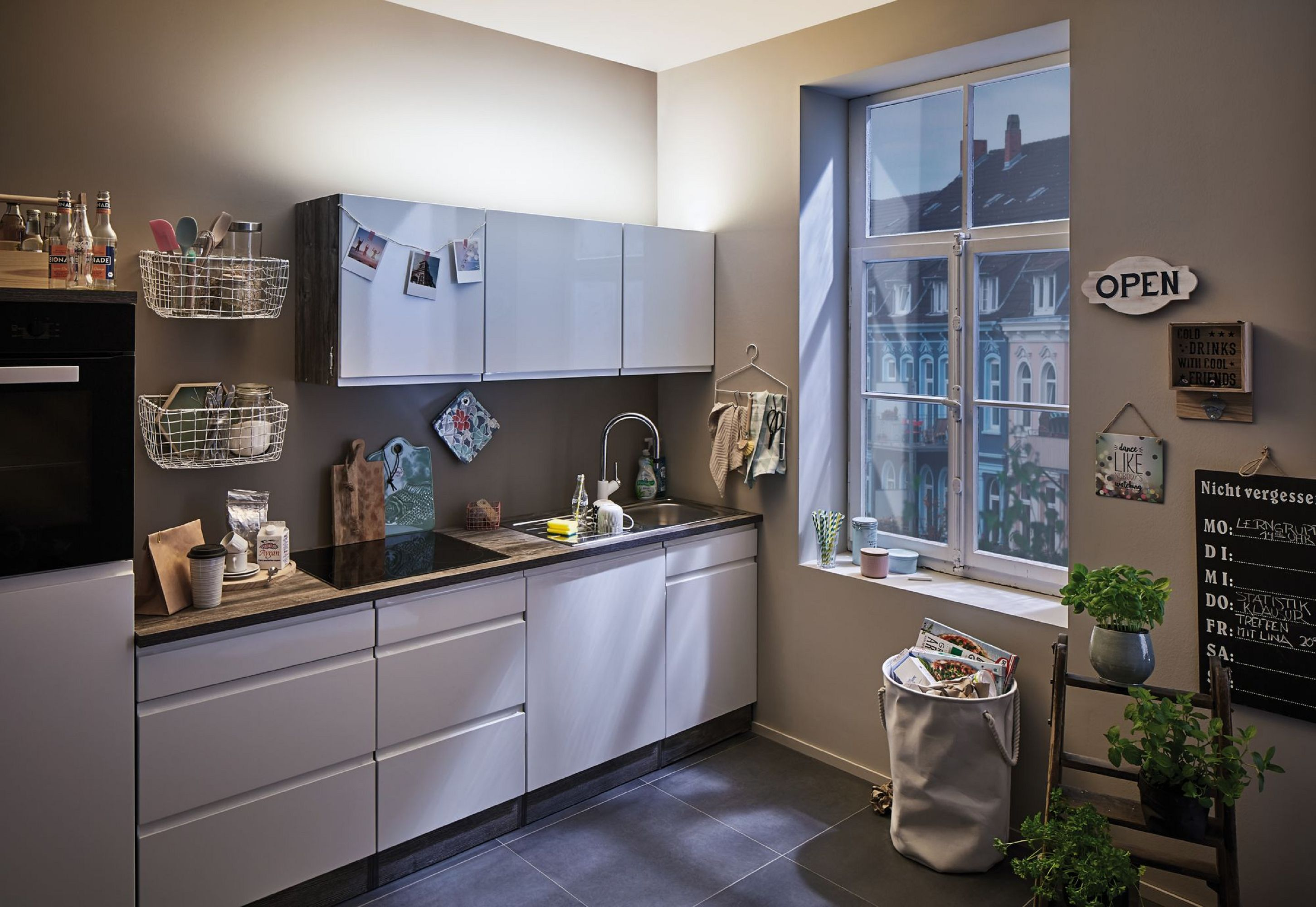 Nowoczesna kuchnia z białymi szafkami, oknem i różnymi dekoracjami. Widoczna jest również tablica i rośliny.