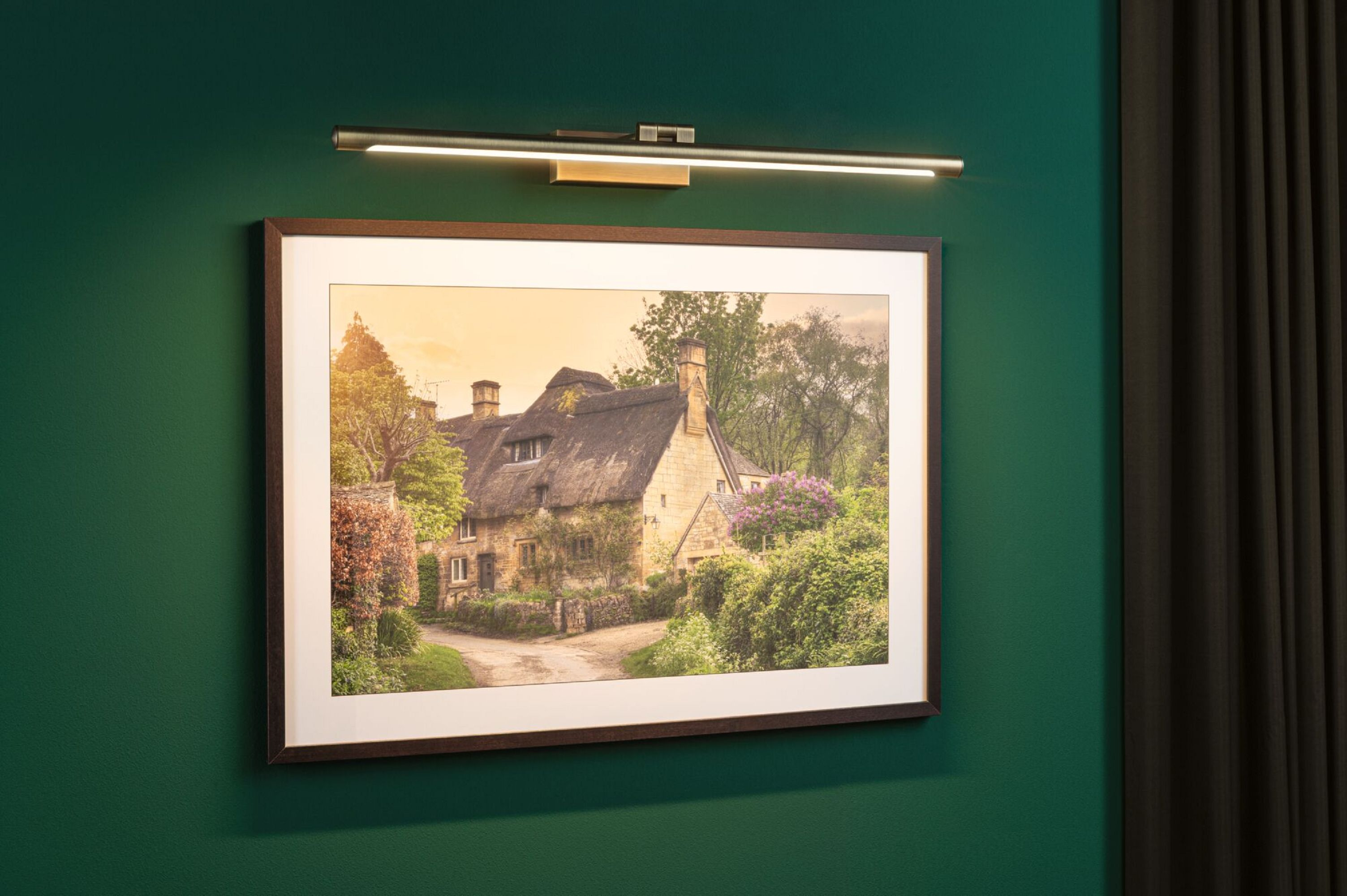 Obraz domku z strzechą, oprawiony i oświetlony przez lampę ścienną, na zielonej ścianie.