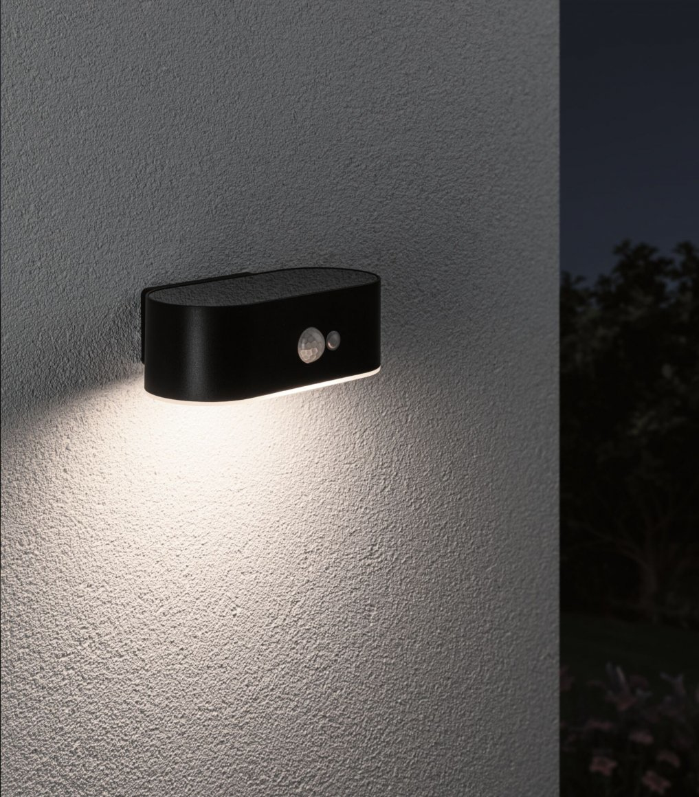 Czarna lampa ścienna z czujnikiem jest zamontowana na teksturowanej szarej ścianie. Jest włączona, rzucając światło w dół. Tło pokazuje liście.