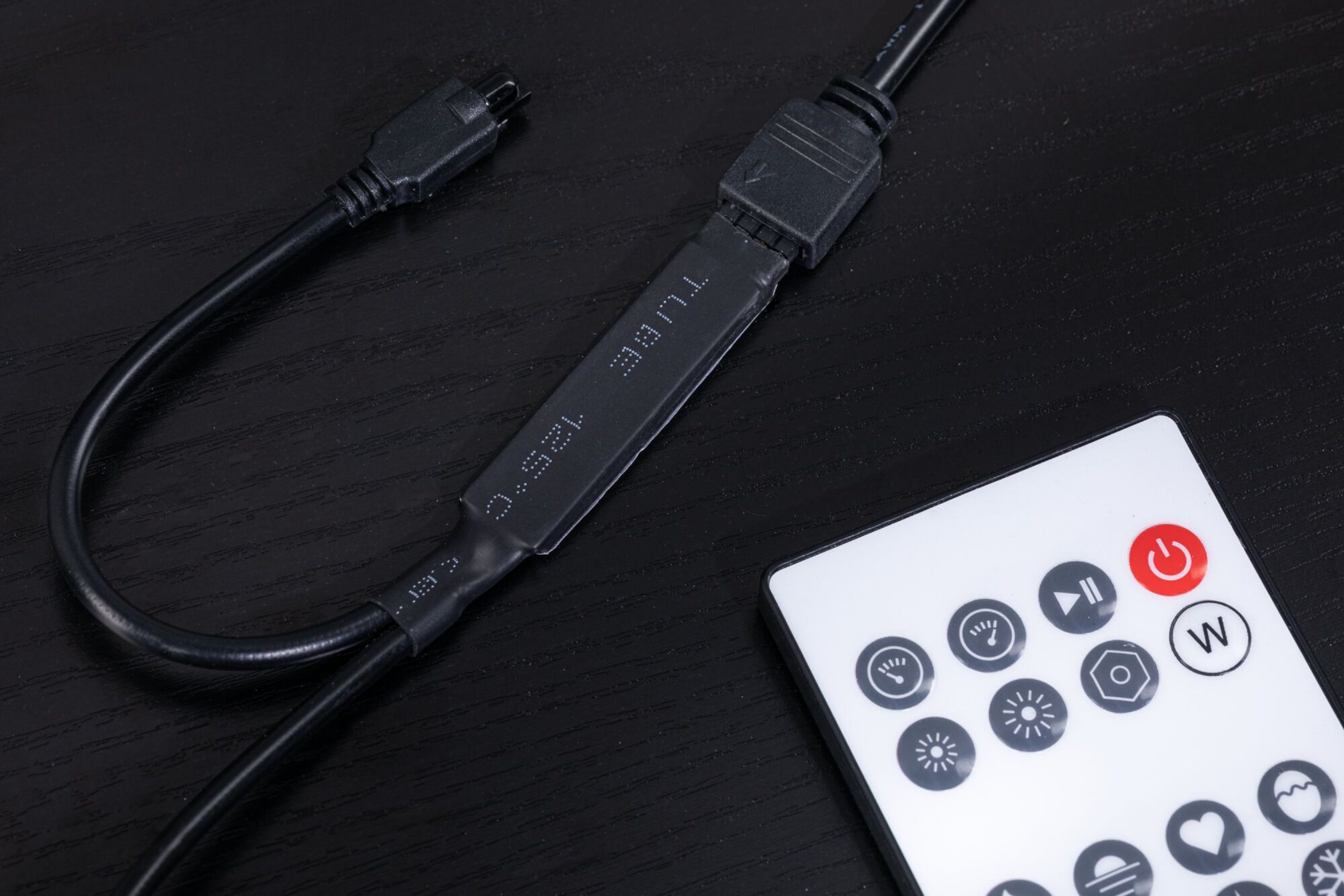 Ein schwarzes Kabel und ein Stecker liegen auf einer schwarzen Holzoberfläche. Eine weiße Fernbedienung liegt daneben.