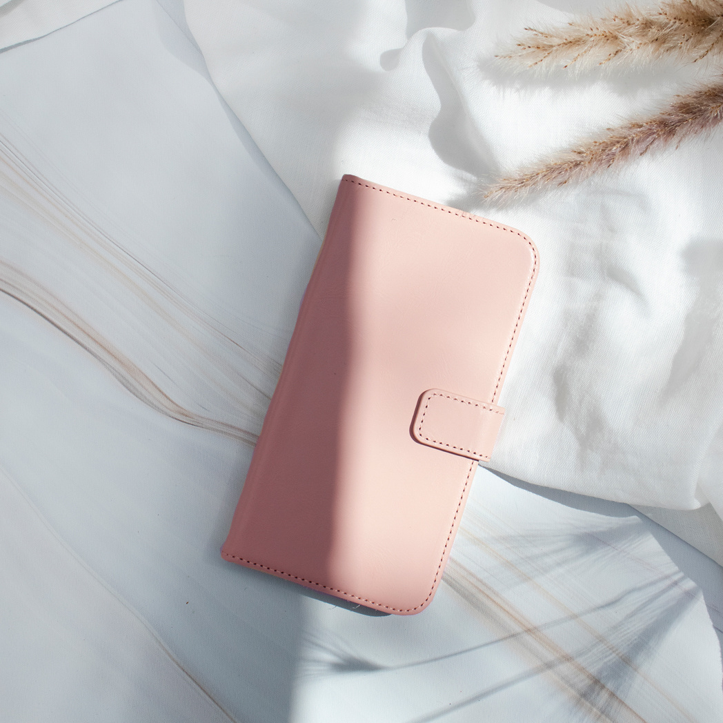 Roze telefoonhoesje op witte stof. Gedroogde planten op de achtergrond. De hoes heeft een magnetische sluiting.