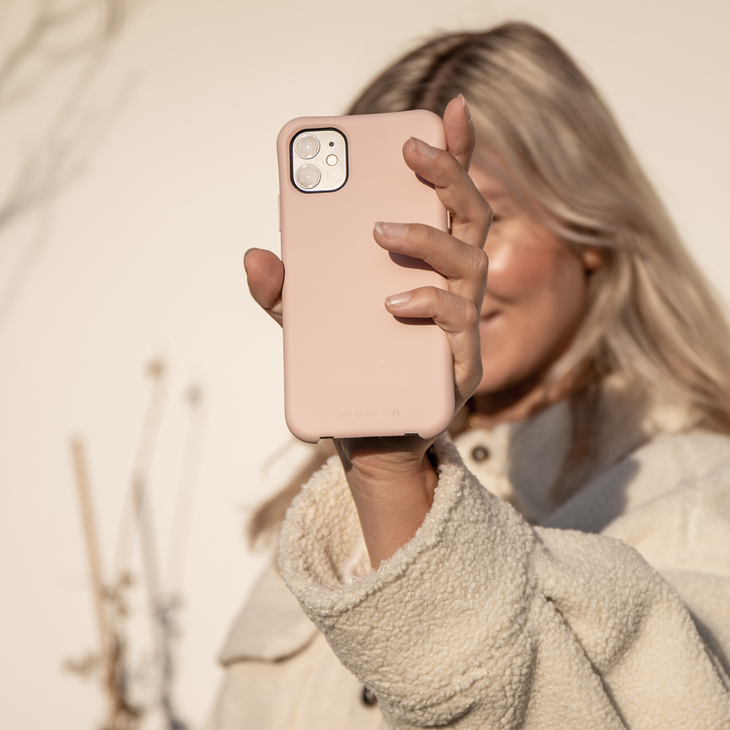 Een vrouw houdt een roze telefoonhoesje voor haar gezicht. Ze draagt een crèmekleurig jasje en heeft blond haar. De achtergrond is beige.