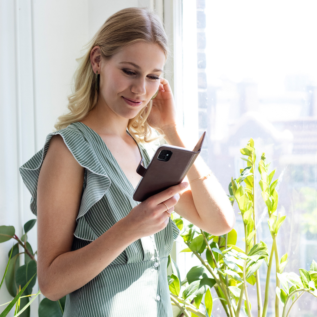 Femme blonde regardant un téléphone dans un étui marron près d'une fenêtre. Elle porte une robe verte. Plantes au premier plan et en arrière-plan.