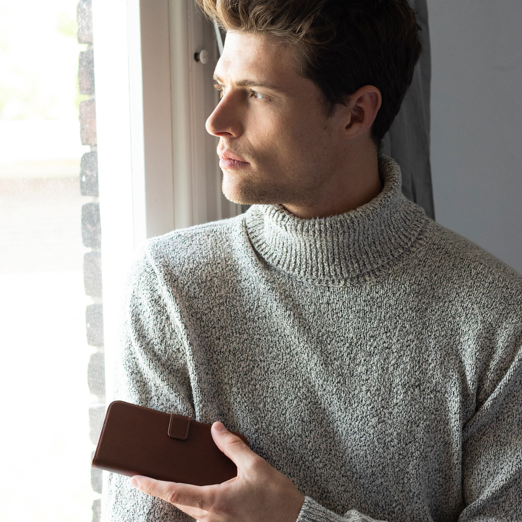 Un homme en col roulé gris regarde par une fenêtre, tenant un portefeuille marron. Le fond montre une fenêtre et un mur clair.