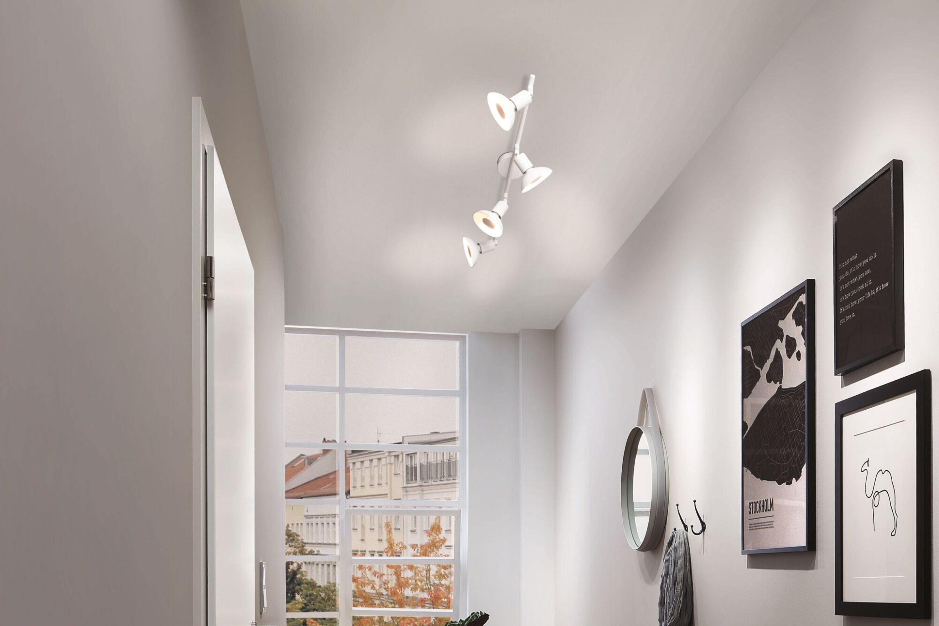 Ein weißer Flur mit Deckenbeleuchtung. An der Wand befinden sich ein Spiegel, Bilder und ein Fenster. Links ist eine Tür.