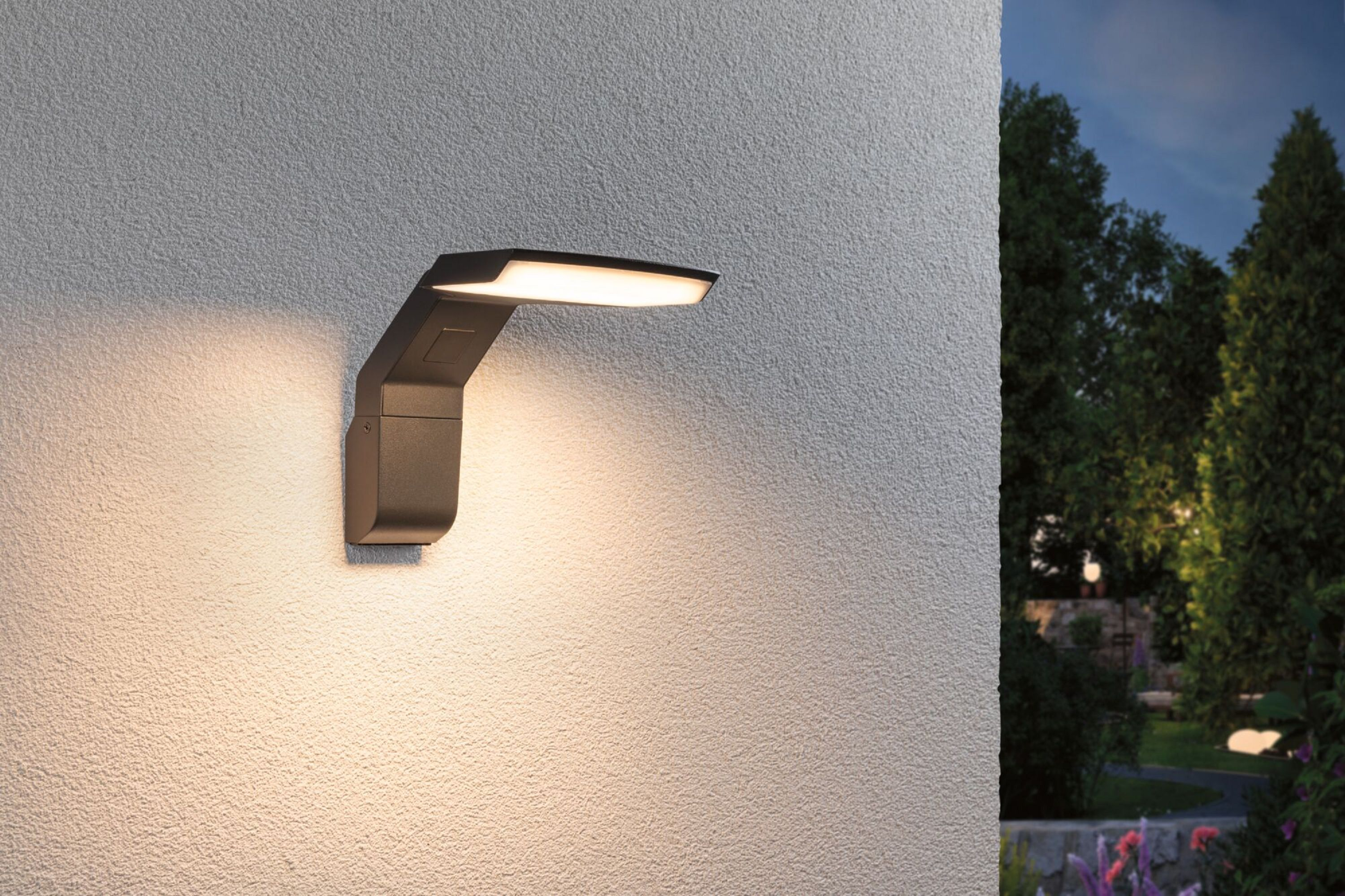 Nowoczesna lampa ścienna emituje światło na teksturowaną ścianę. Drzewa są w tle.