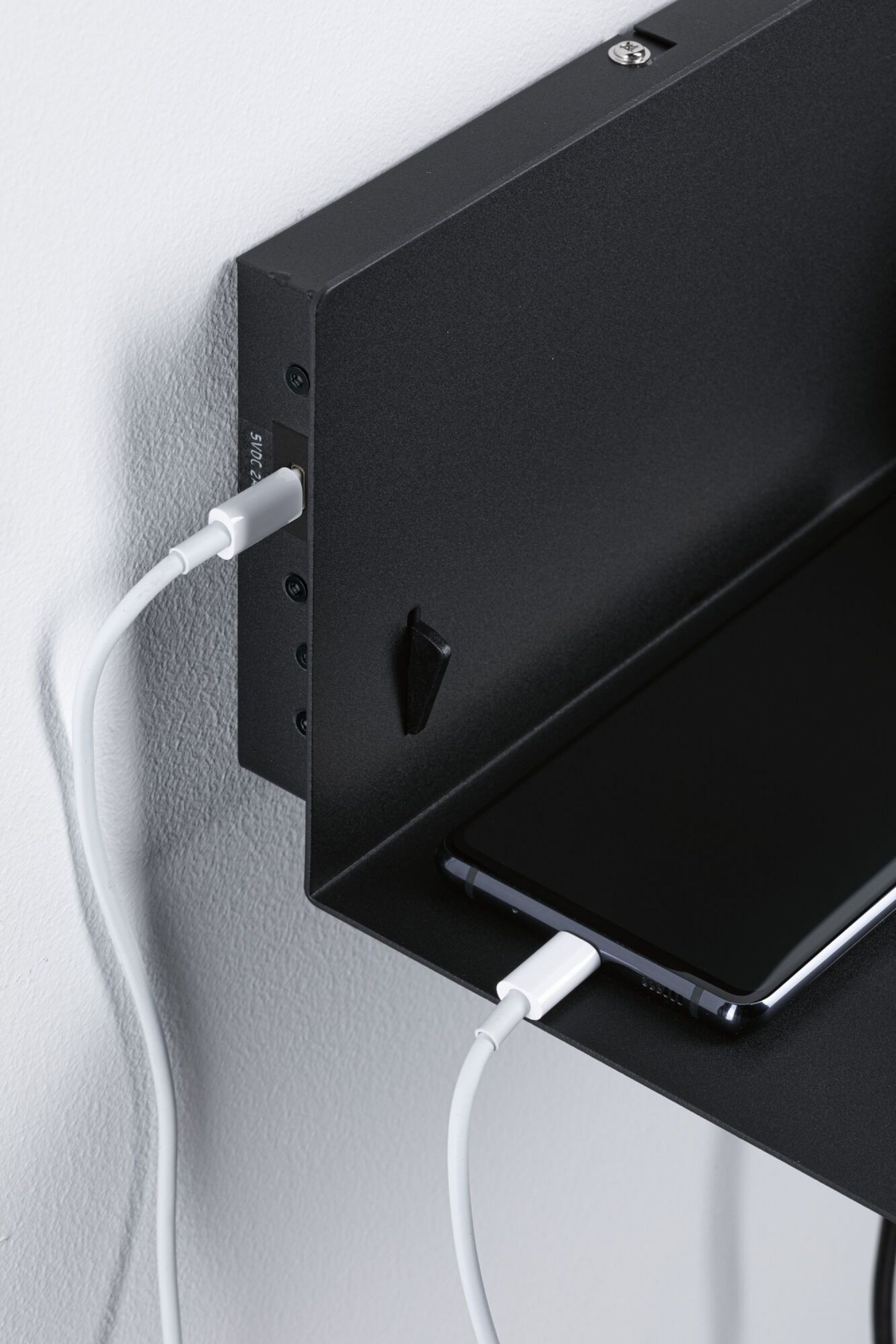 Czarna półka z ładującym się telefonem. Biały kabel podłączony do telefonu. Czarny uchwyt ścienny.