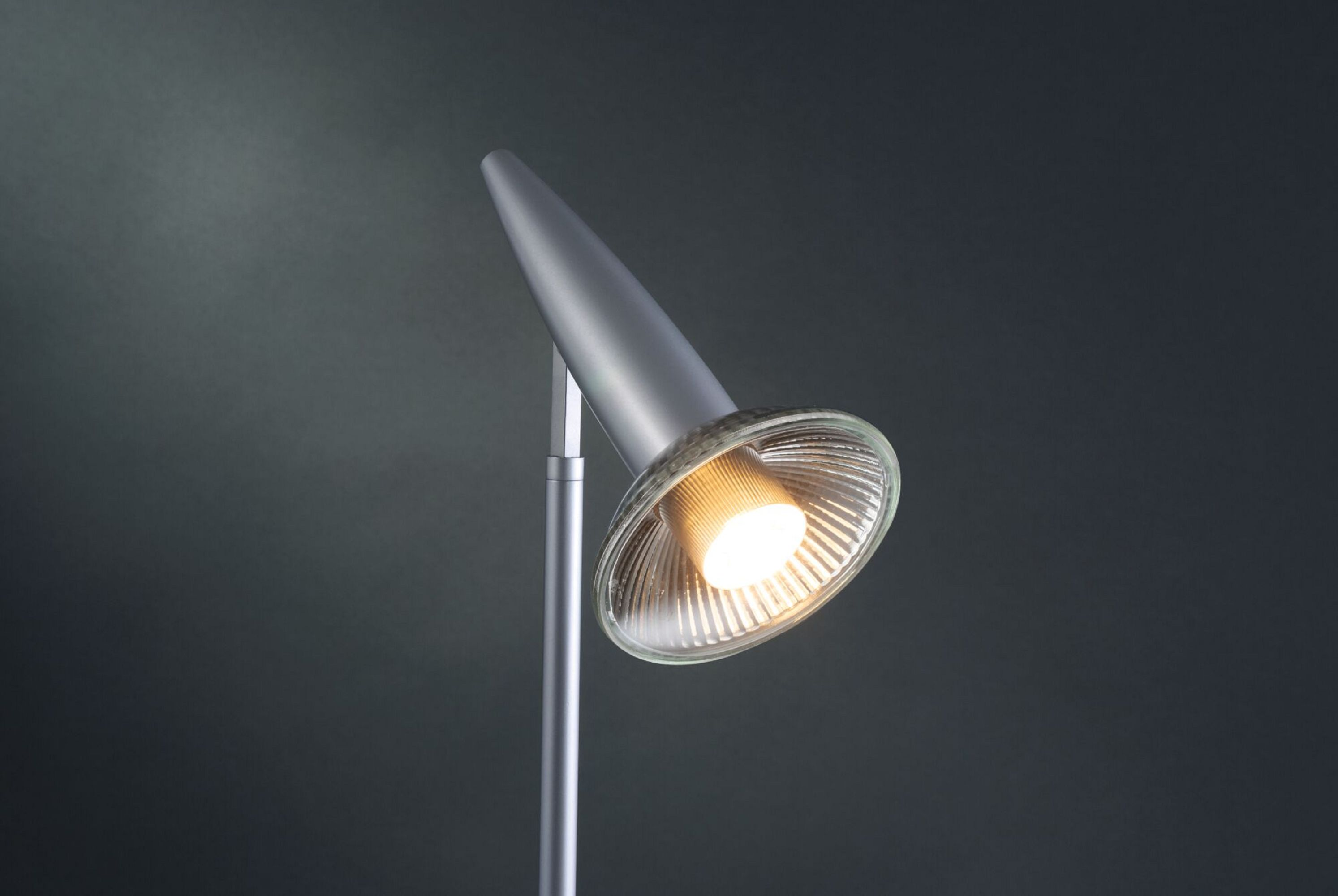 Srebrna lampa biurkowa z stożkową głowicą i przezroczystą, żebrowaną soczewką, na ciemnoszarym tle.