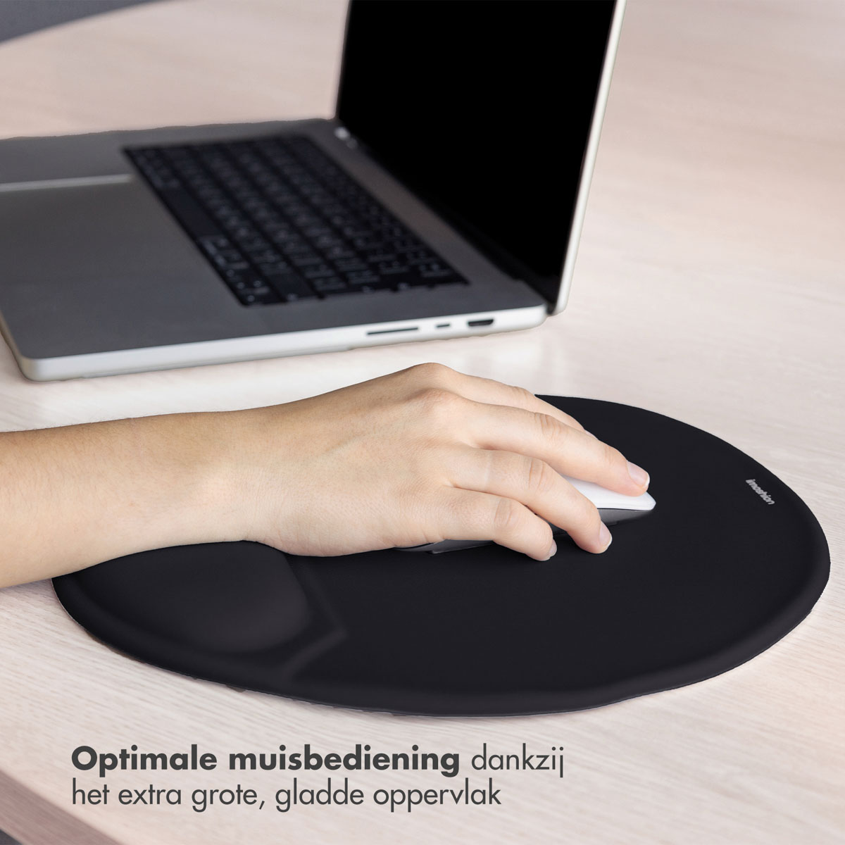 A hand uses a mouse on a black mousepad with a wrist rest. A laptop is in the background.