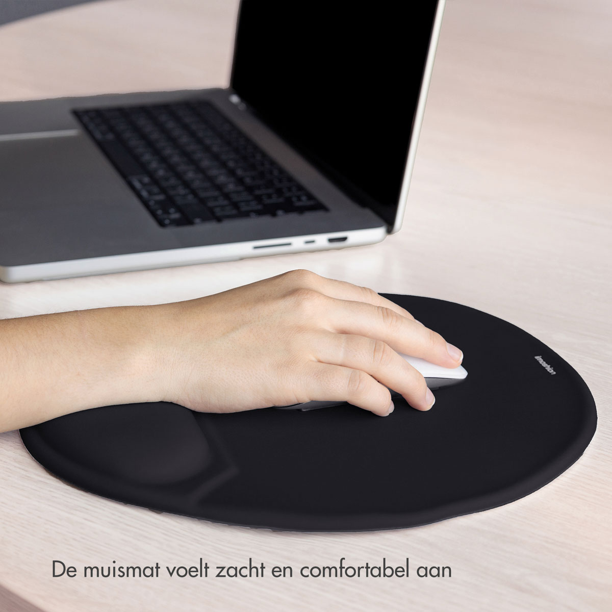 A hand operates a white mouse on a black mousepad. A laptop sits in the background. The scene is on a light wooden table.