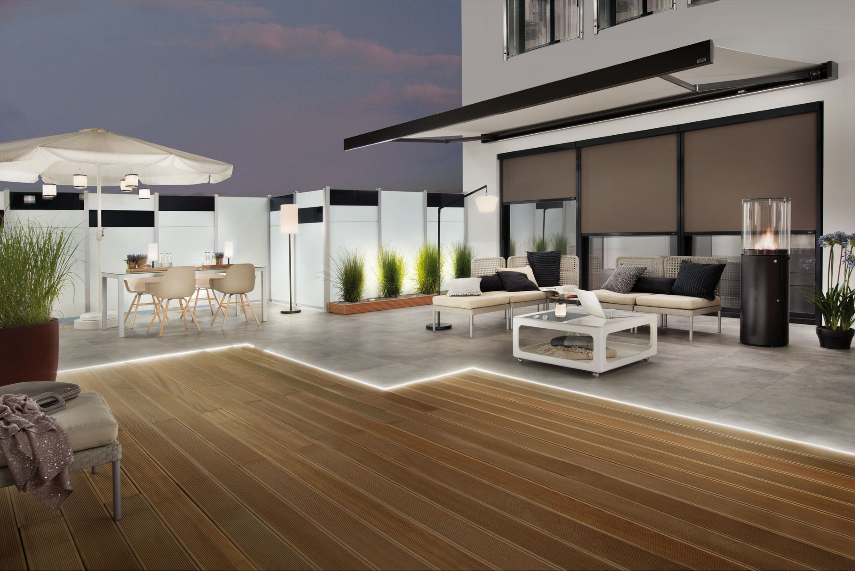 Patio na świeżym powietrzu z białym parasolem, stołem, krzesłami i sofą. Obecny jest również kominek i rośliny. Drewniany pokład.