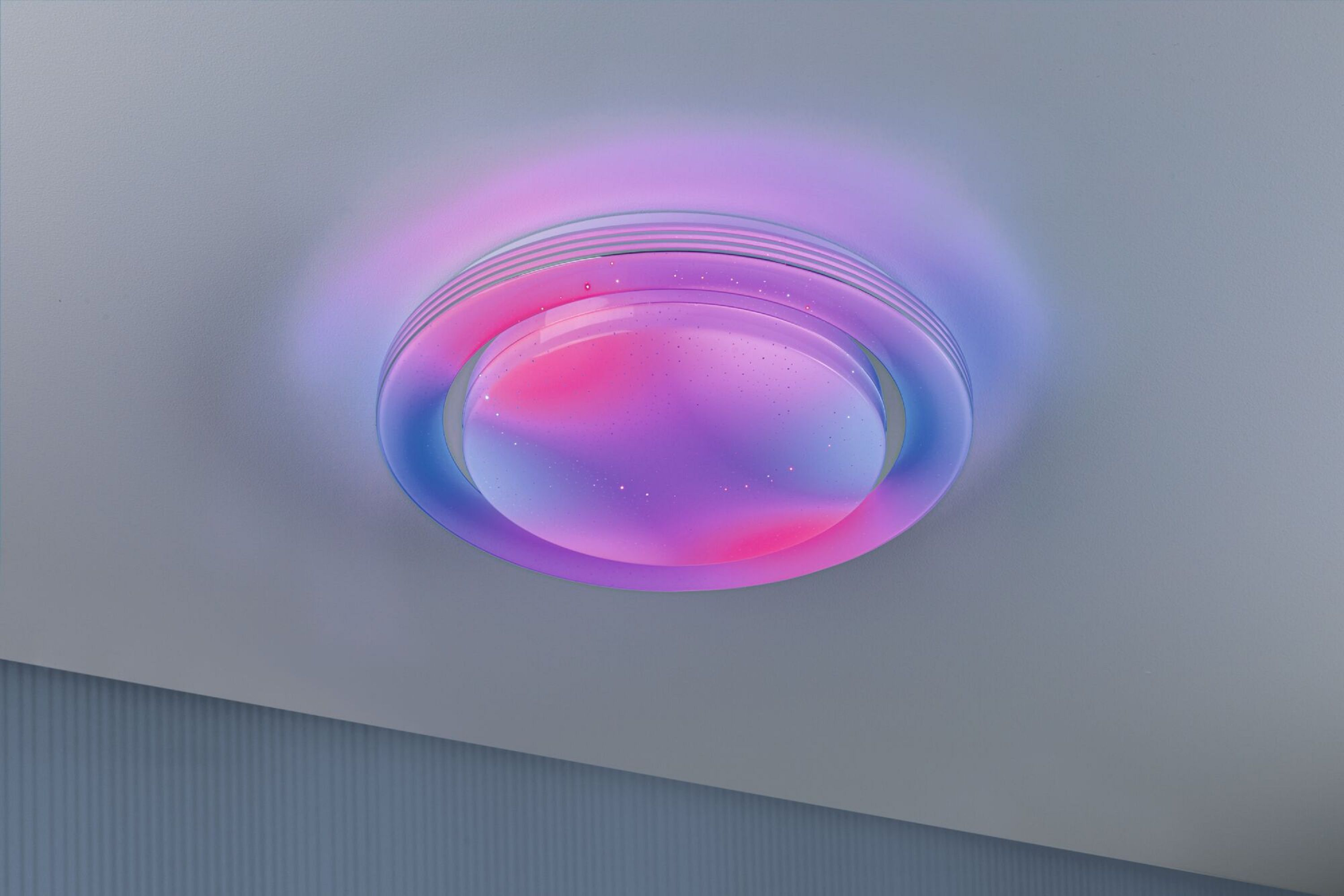 Un apparecchio di illuminazione a soffitto con luce rosa e blu, su un soffitto grigio. Il design è circolare.