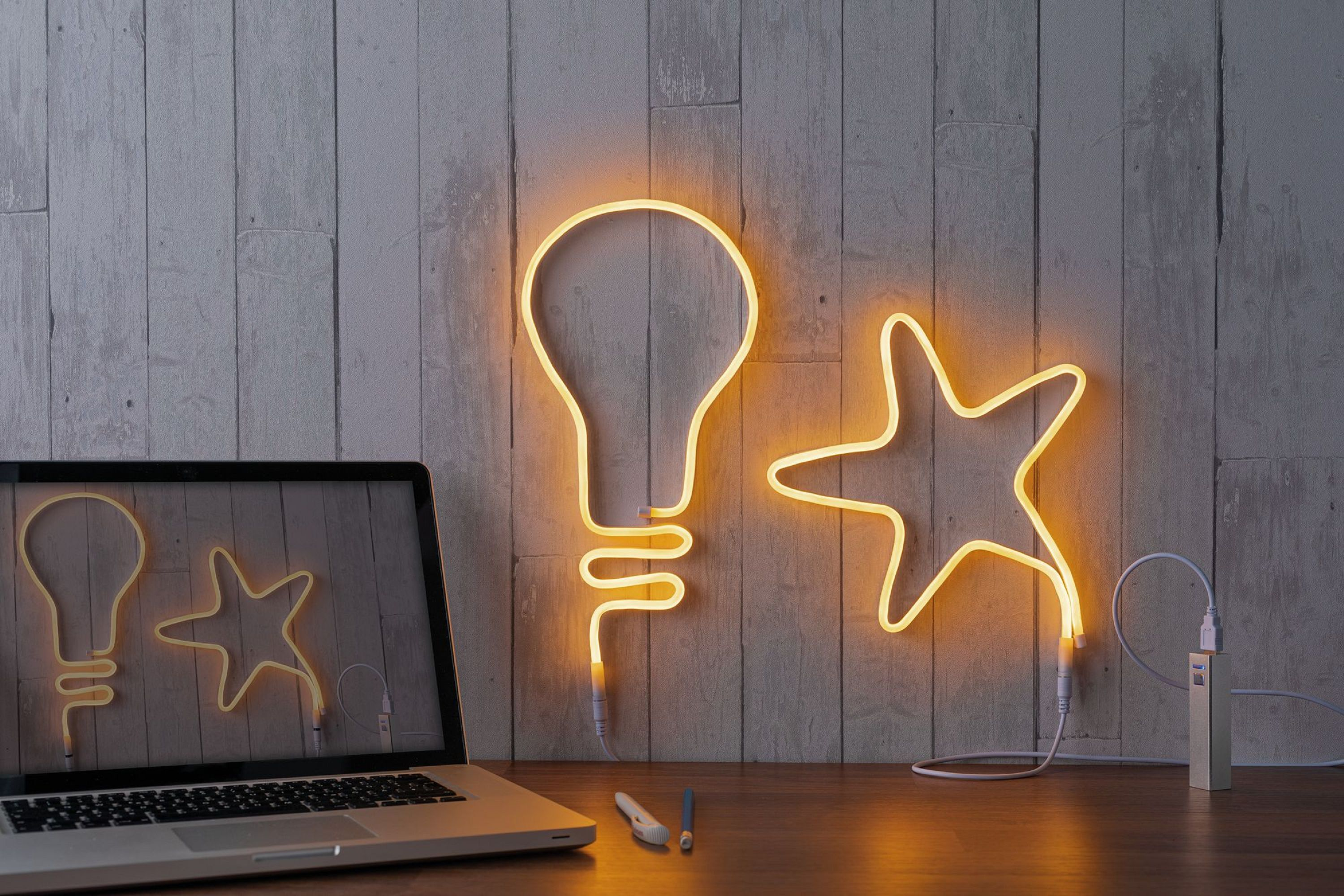 Un laptop, una lampadina e una stella al neon, su una superficie di legno, contro un muro di legno grigio.