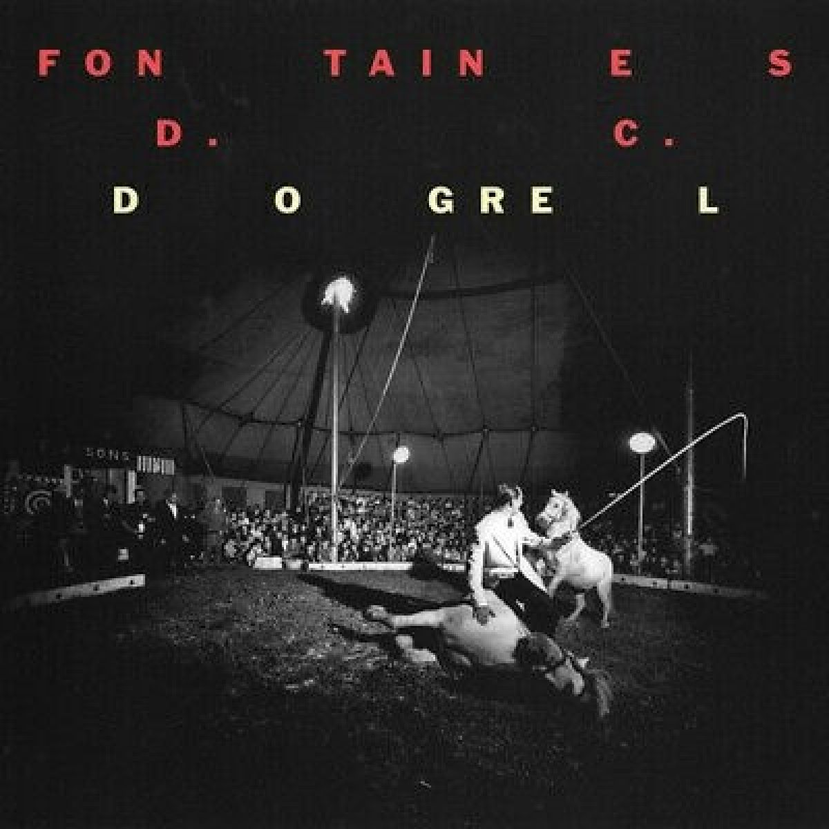 Scena circense con artisti, cavalli e pubblico. Un uomo è vicino a un cavallo. La scena è buia, illuminata da riflettori.