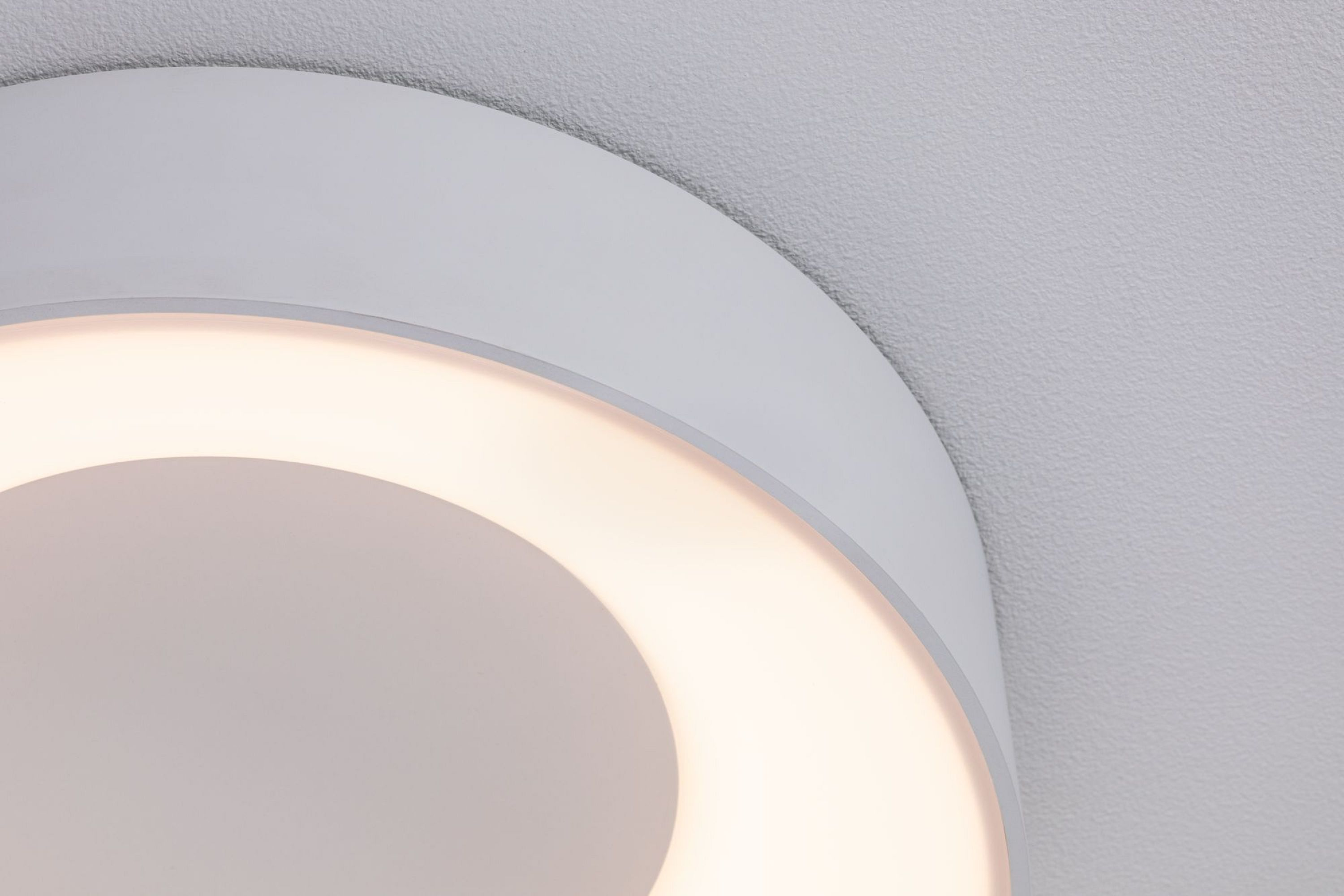 Primo piano di un apparecchio di illuminazione bianco, circolare, con un anello interno luminoso e caldo. Contro un soffitto bianco.