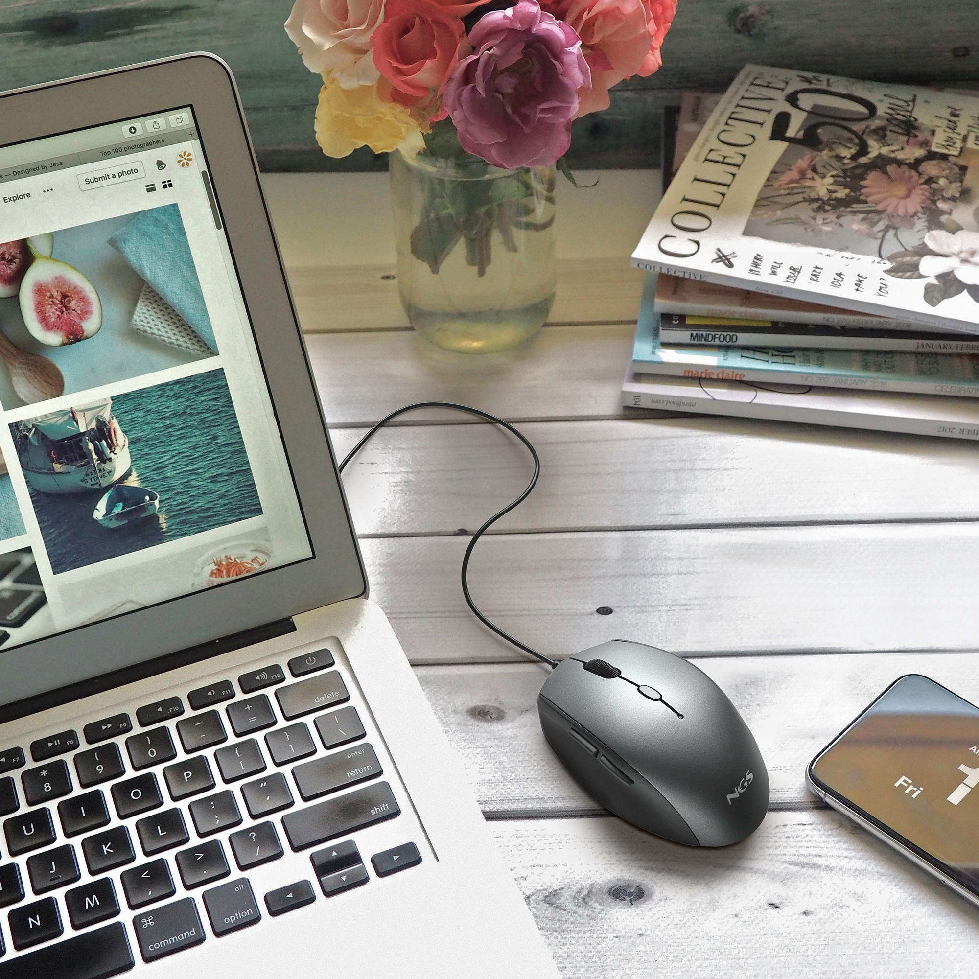 Laptop, mysz, telefon na drewnianym stole. Obecne są również kwiaty w wazonie i czasopisma.