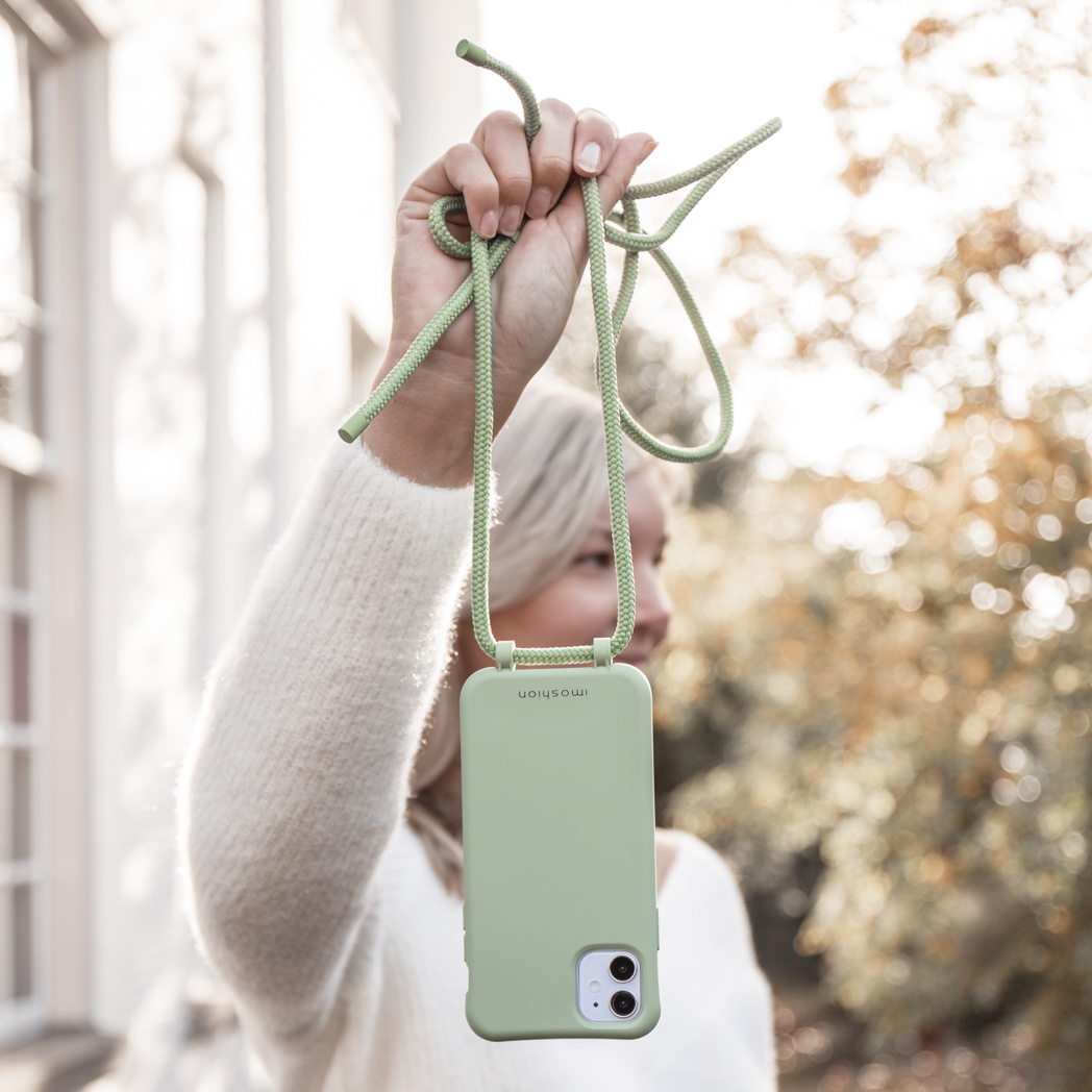Een vrouw houdt een lichtgroene telefoonhoes met een bijpassend koord vast. De achtergrond is wazig met bomen en zonlicht.