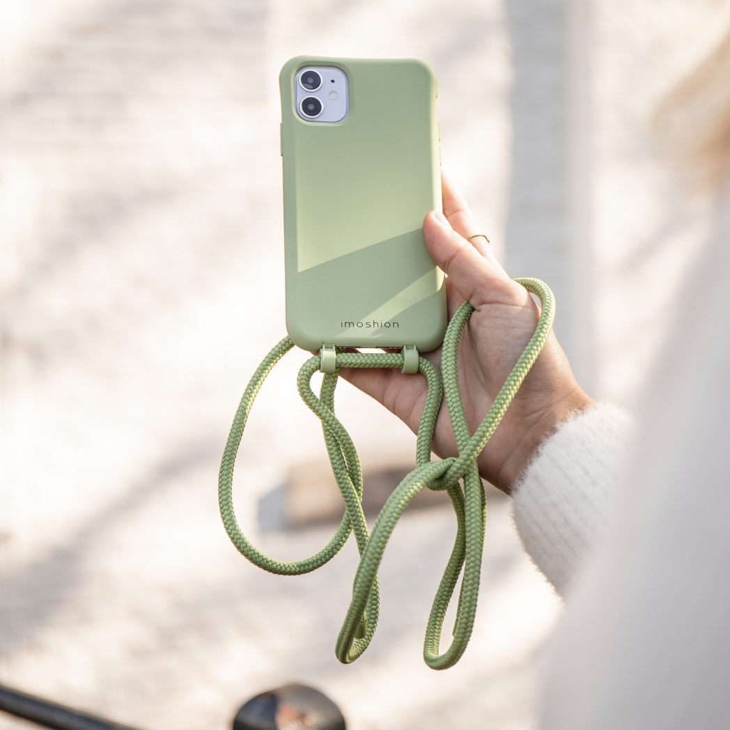Een hand houdt een groene telefoonhoes met een bijpassende riem vast. De hoes is bevestigd aan de riem, die naar beneden hangt. De achtergrond is wazig.