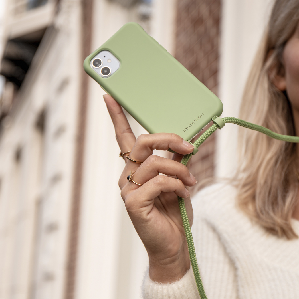 Een vrouw houdt een groene telefoon vast met een bijpassende band. Ze draagt ringen en een witte trui. Gebouwen zijn zichtbaar op de achtergrond.