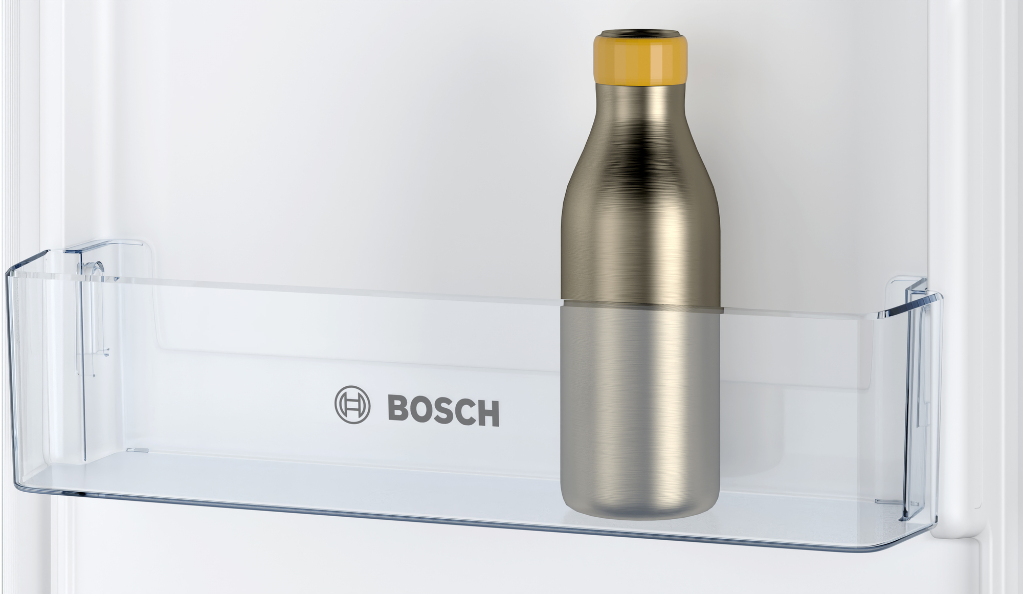 Metalowa butelka ze złotą nakrętką stoi na przezroczystej plastikowej półce w białej lodówce. Widoczne jest logo Bosch.