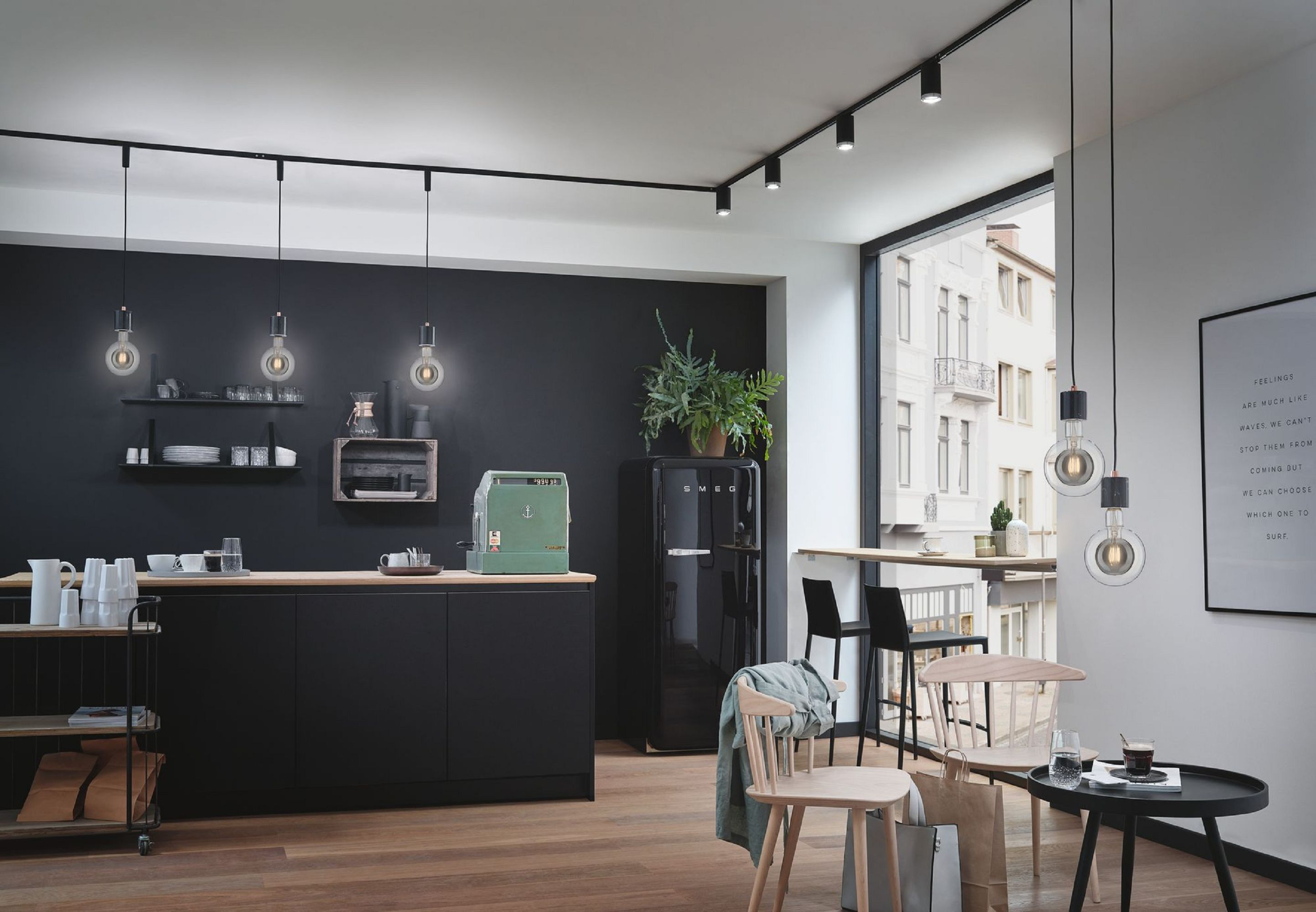 Moderne Küche mit schwarzen Schränken, einer grünen Kaffeemaschine, einem schwarzen Kühlschrank, Holzboden und Hängelampen. Ein Poster an der Wand.