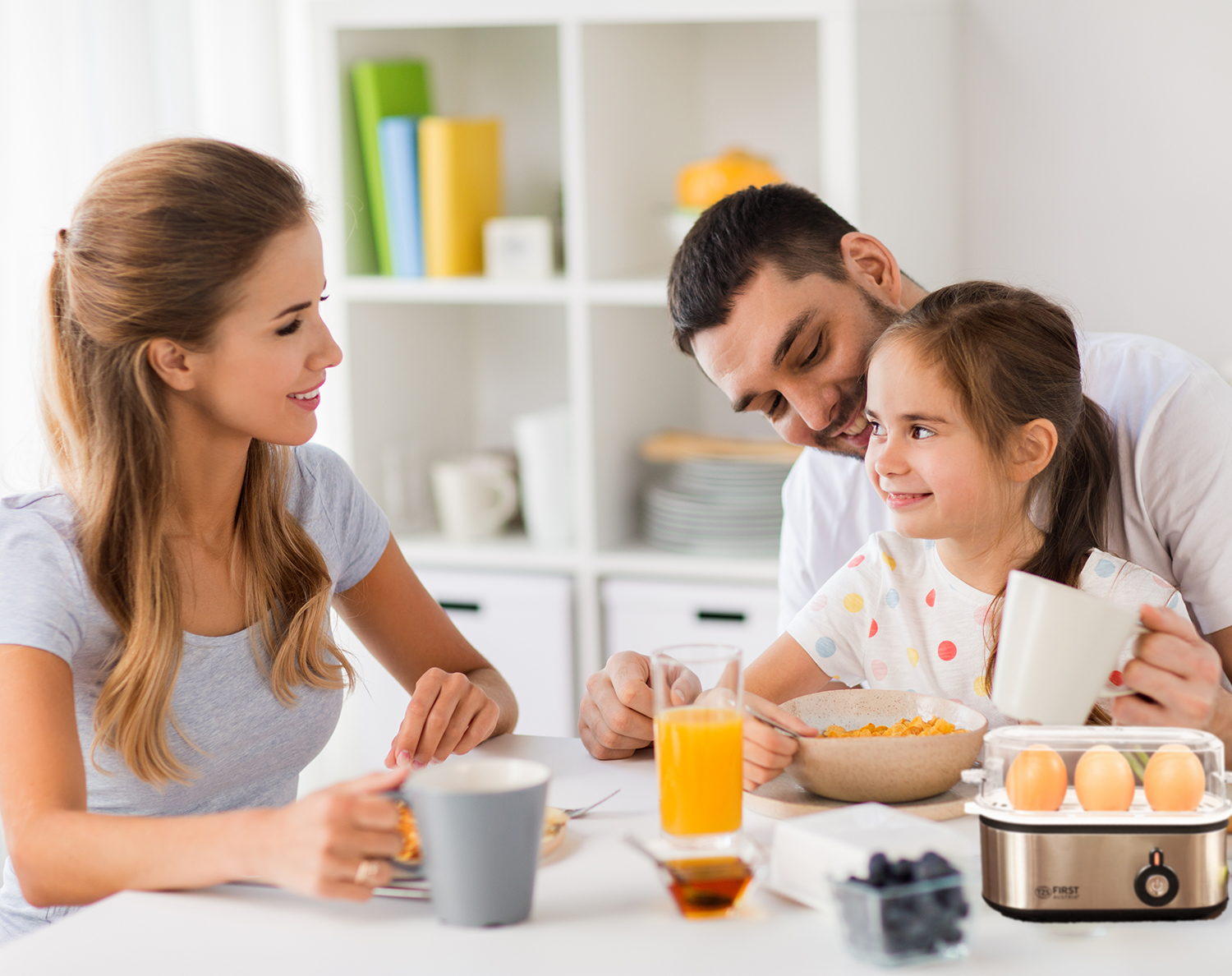 Eine Familie frühstückt gemeinsam. Eine Frau, ein Mann und ein Kind sitzen an einem Tisch mit Essen und Getränken. Ein Eierkocher steht auf dem Tisch.