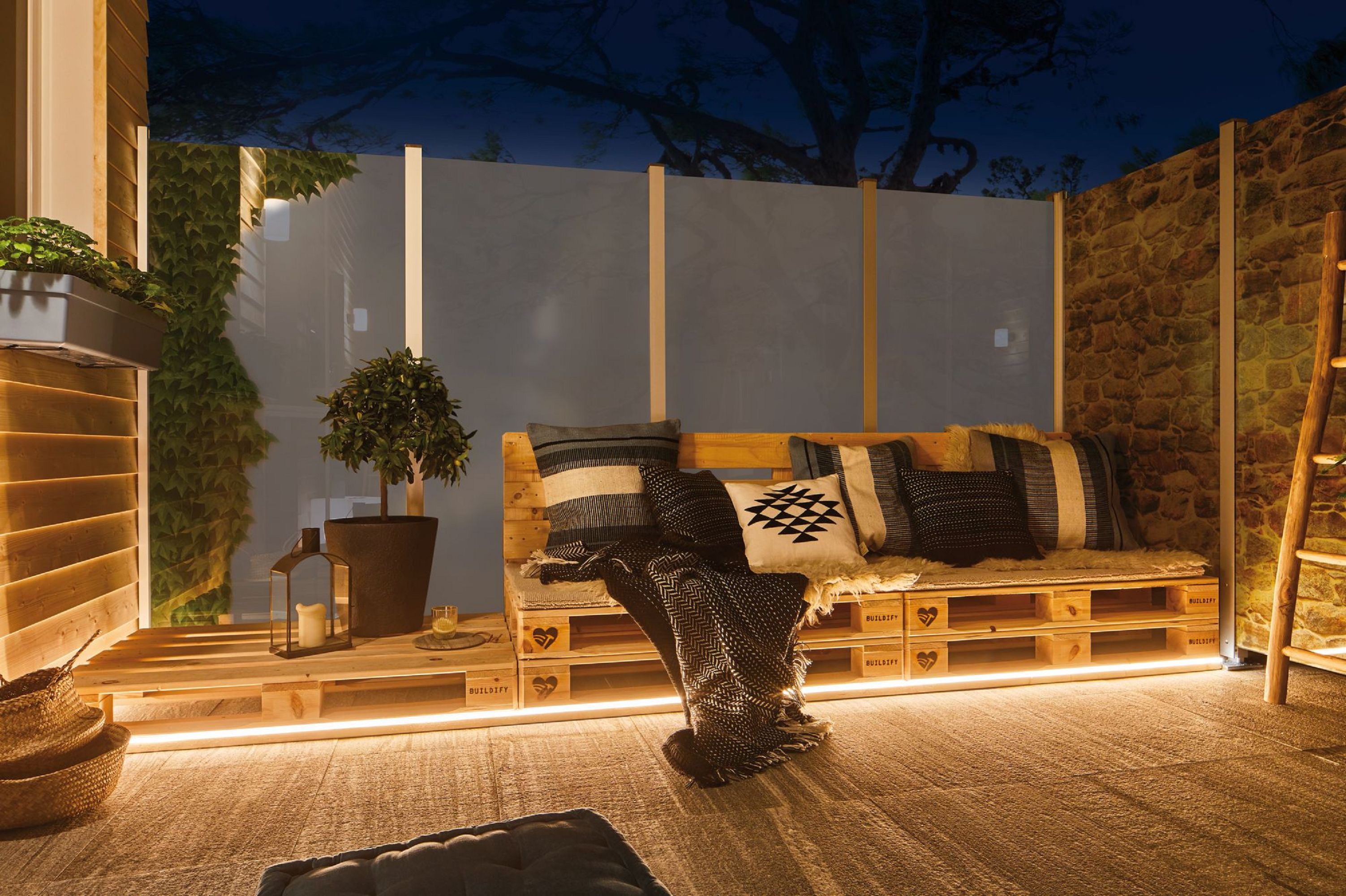 Outdoor-Sitzbereich bei Nacht. Palettensofa mit Kissen und Decken. Laterne und Topfpflanze. Hintergrund aus Steinmauer.