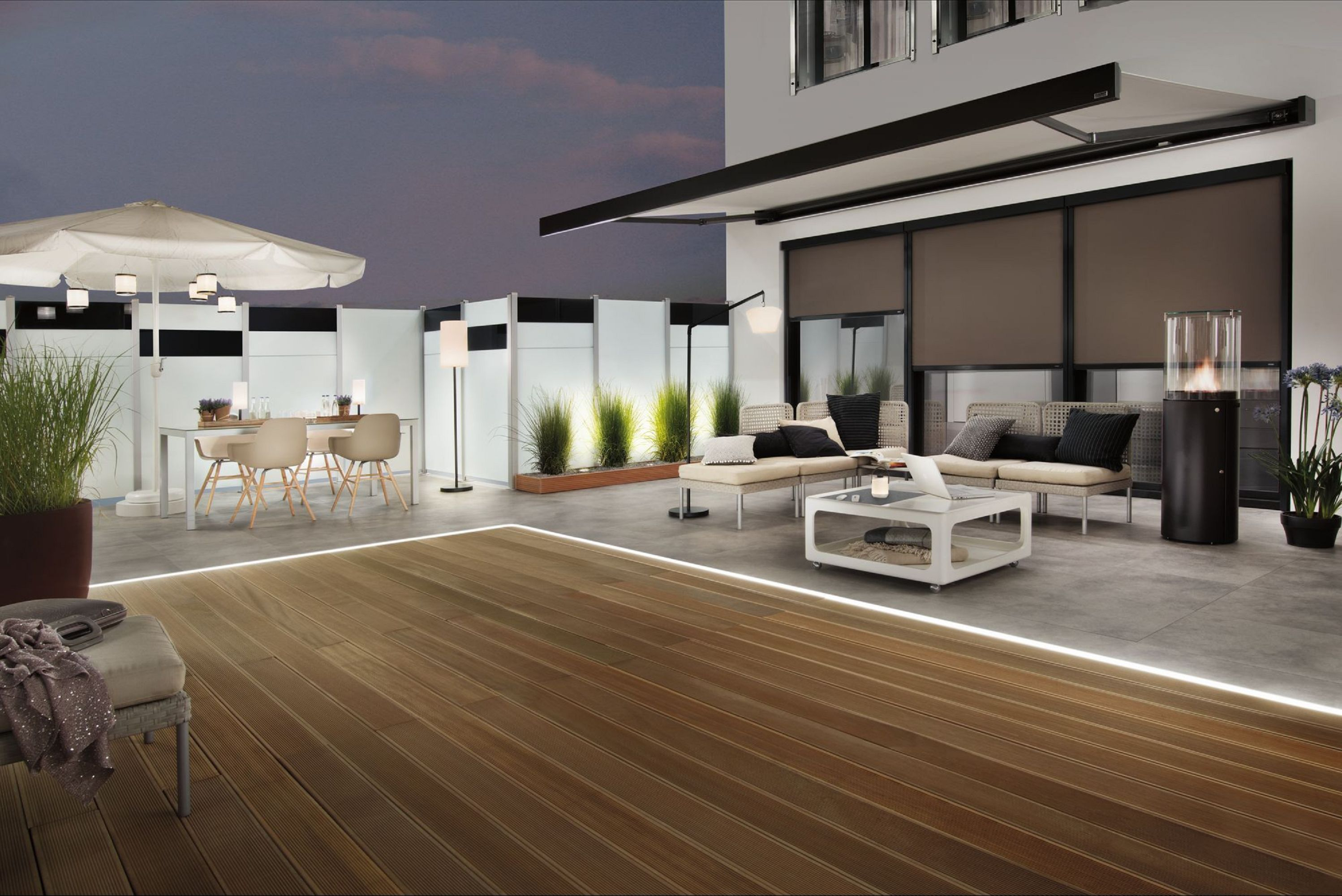 Terrasse im Freien mit Holzdeck, Tischen, Stühlen und Sofas. Pflanzen und Lampen sind sichtbar. Der Himmel ist Dämmerung.