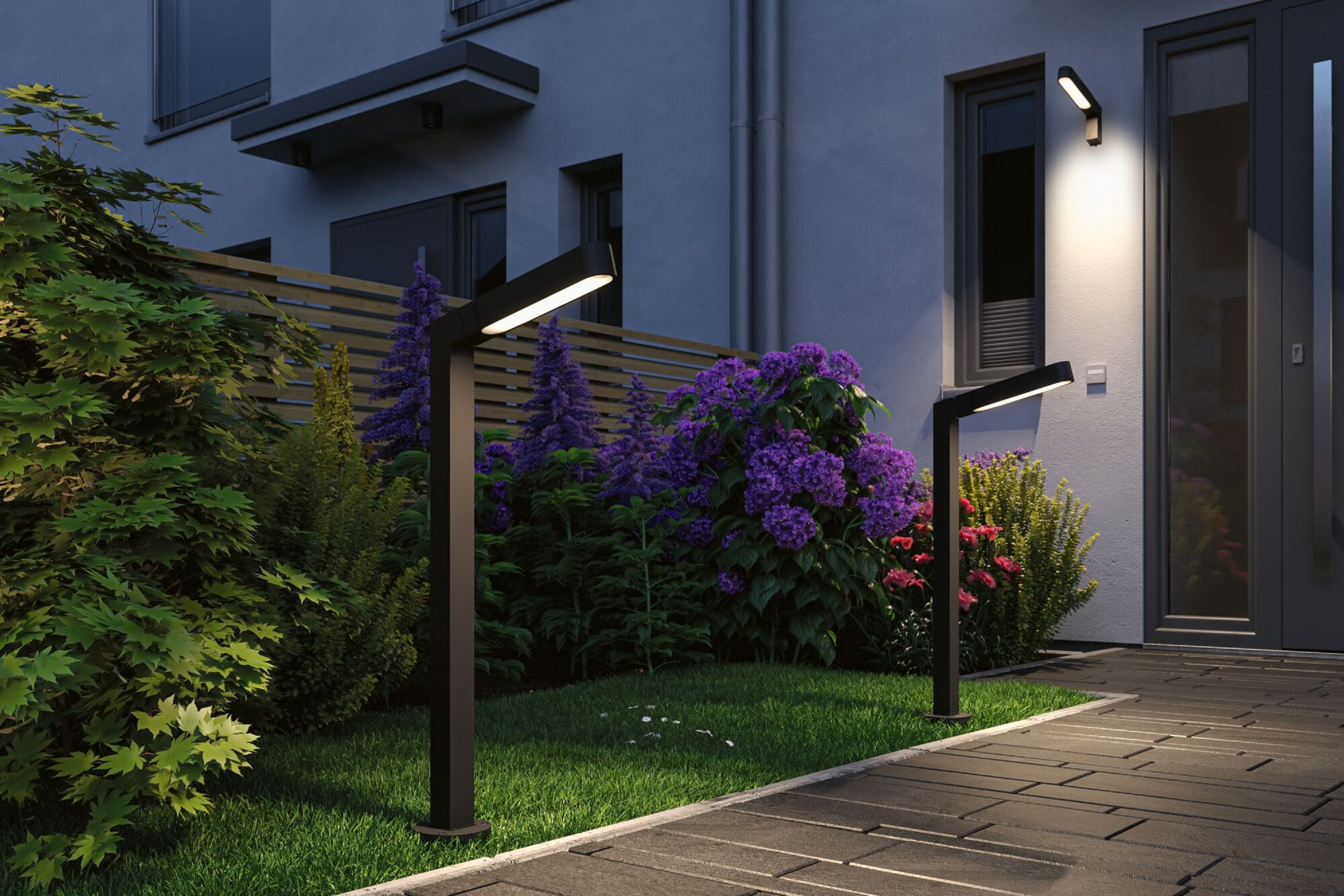 Außenansicht in der Abenddämmerung. Moderne Lampen beleuchten einen Weg. Lila Blumen und grüne Büsche. Ein Gebäude im Hintergrund.