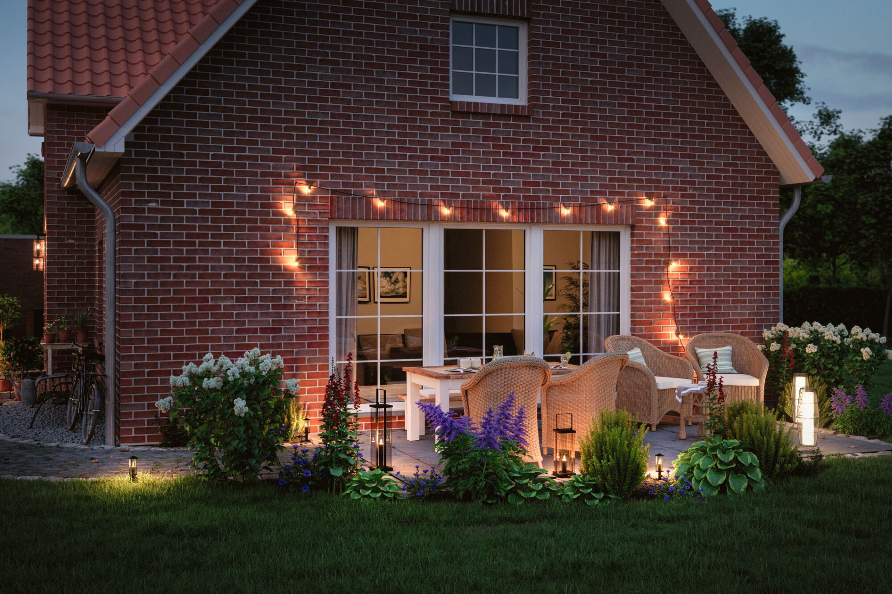 Außenszene eines Backsteinhauses mit Terrassenmöbeln. Lichterketten sind an. Blumen und Büsche umgeben die Terrasse.