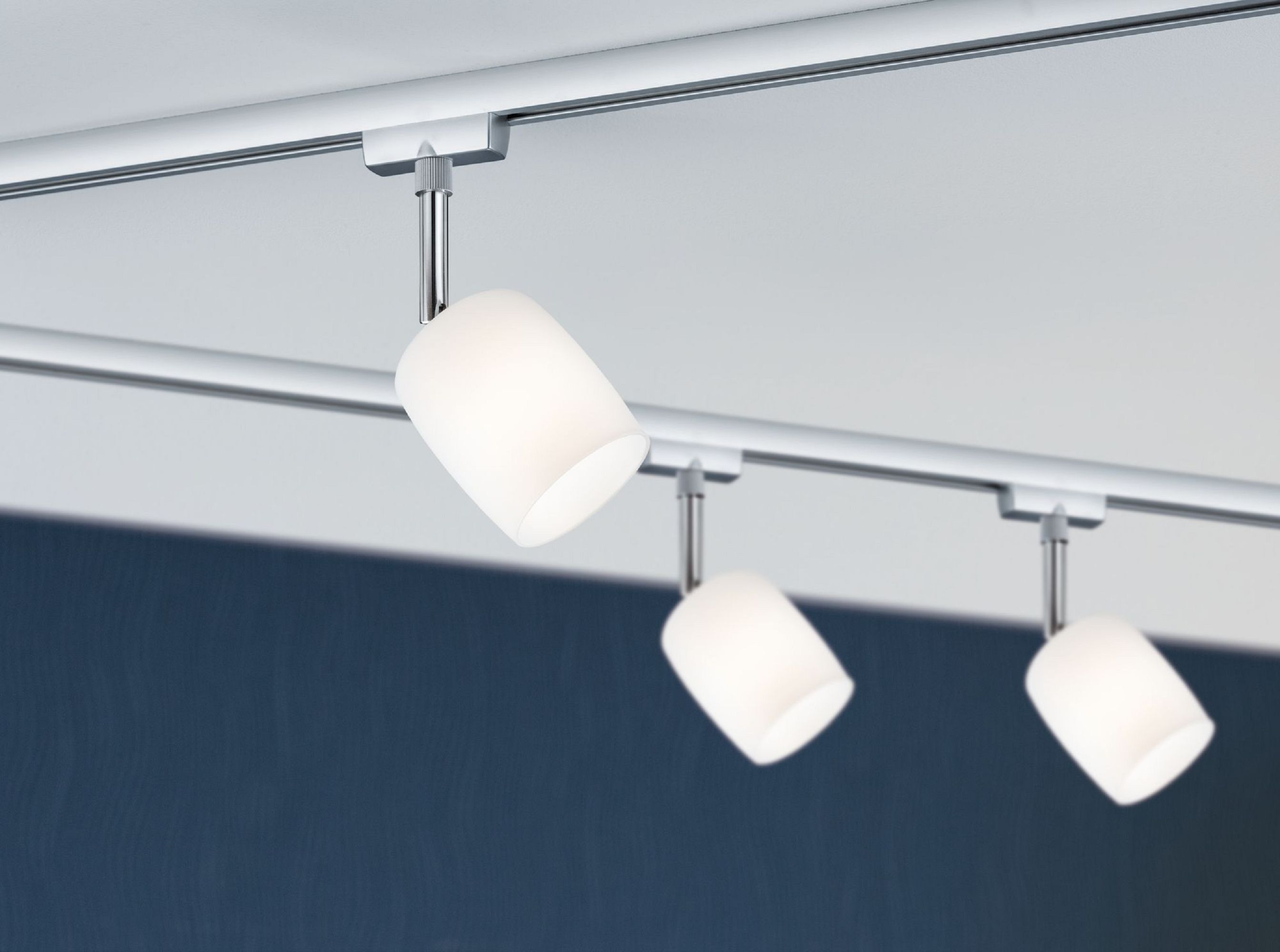 Drei weiße Strahler an einer silbernen Schiene vor weißer Decke und dunkelblauer Wand.