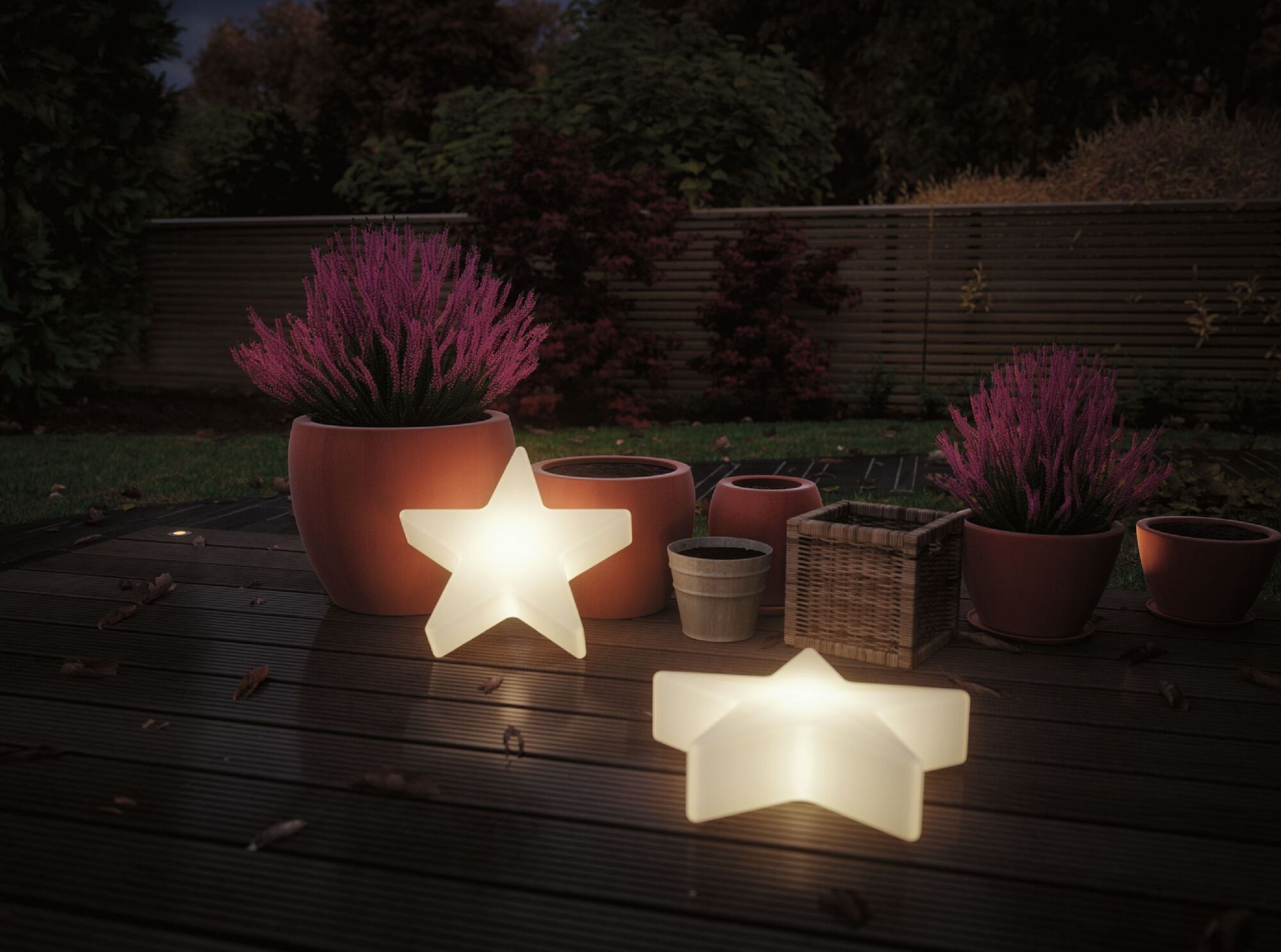 Nächtliche Außenszene. Zwei leuchtende sternförmige Lichter, Töpfe mit Pflanzen und eine Holzterrasse. Dunkler Hintergrund mit Bäumen.