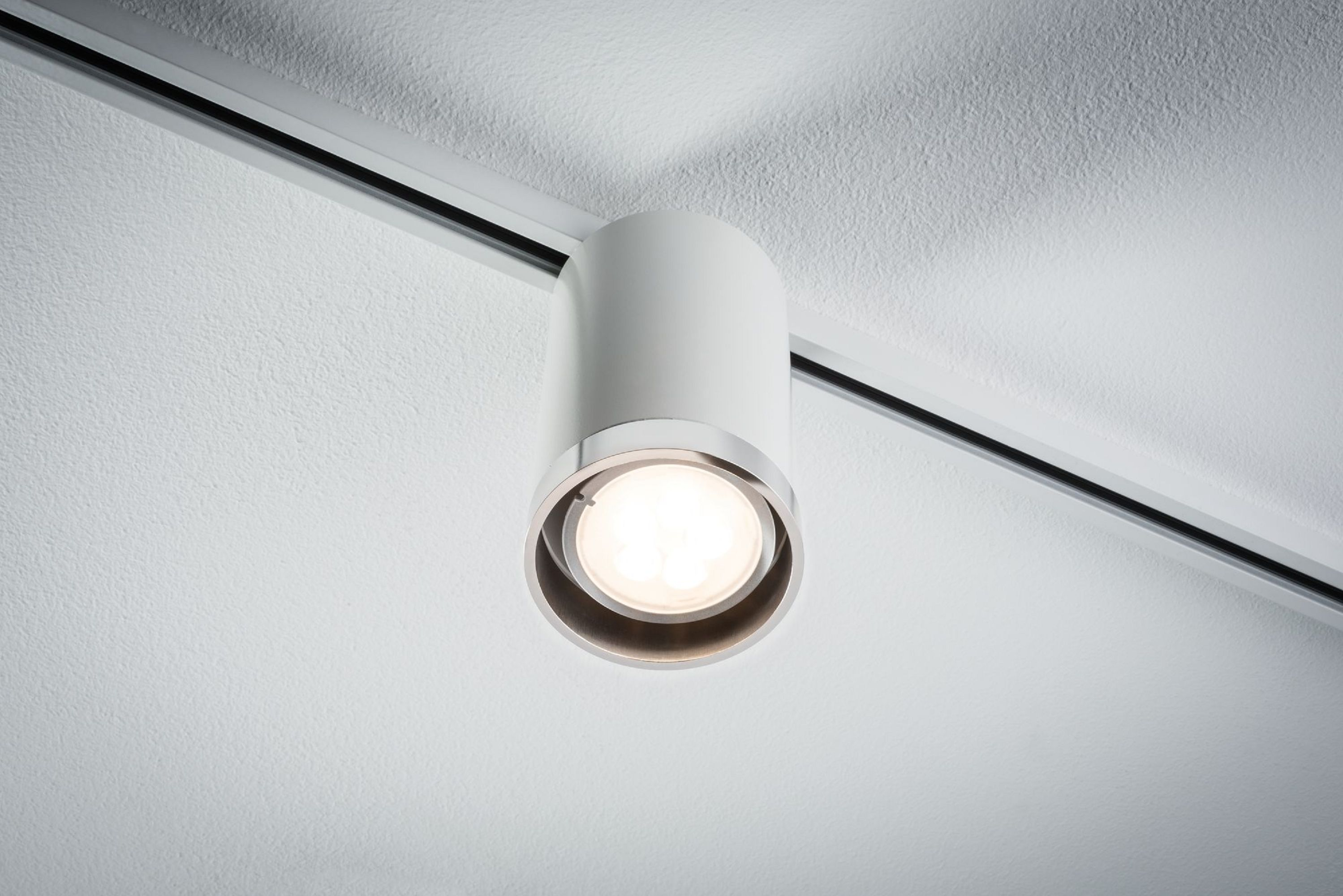 Ein weißer zylindrischer Strahler hängt von einer weißen Decke. Er leuchtet nach unten. Eine schwarze Schiene verläuft über die Decke.