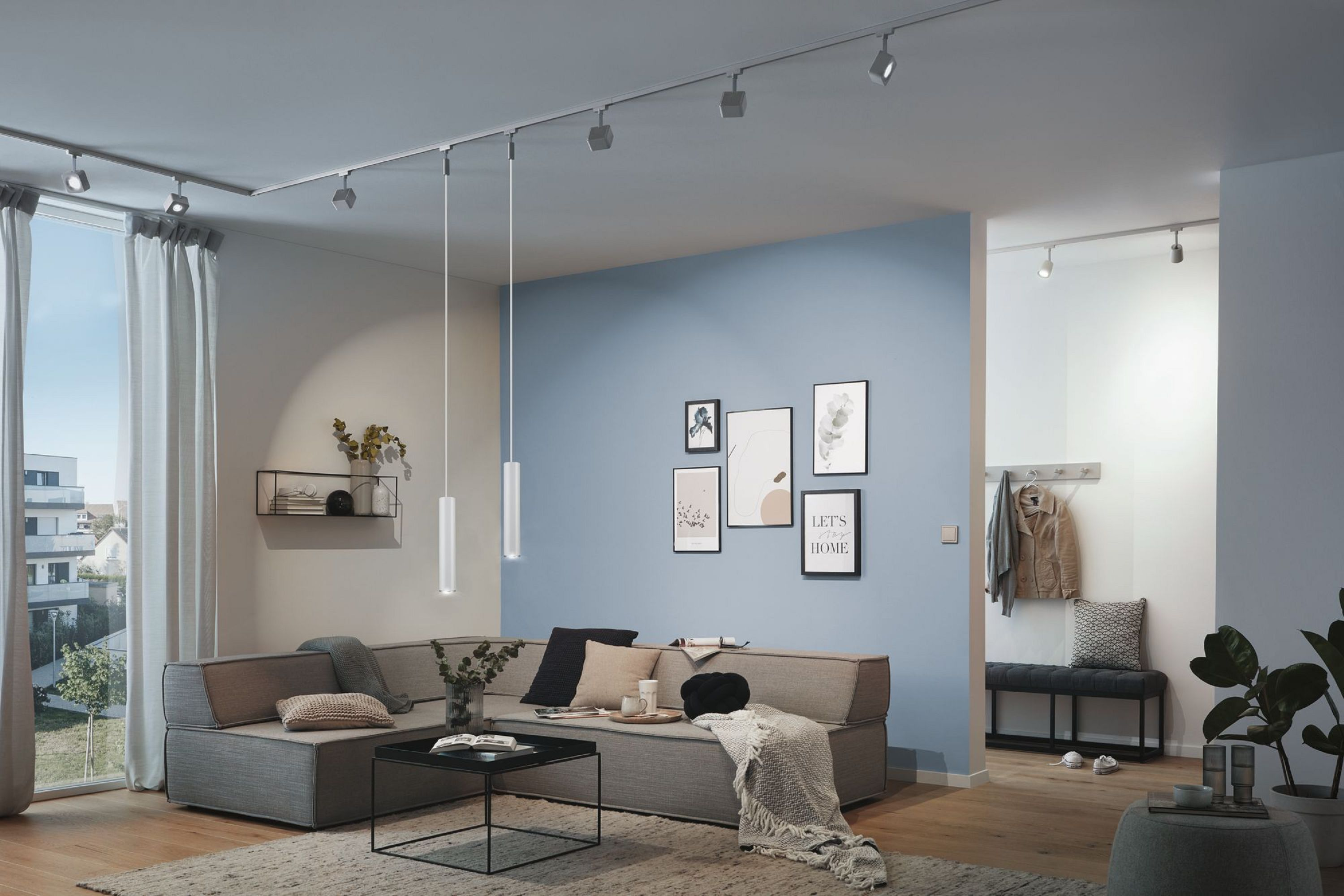 Ein modernes Wohnzimmer mit Sofa, Bildern, Schienenbeleuchtung und Teppich. Rechts ist ein Flur zu sehen.