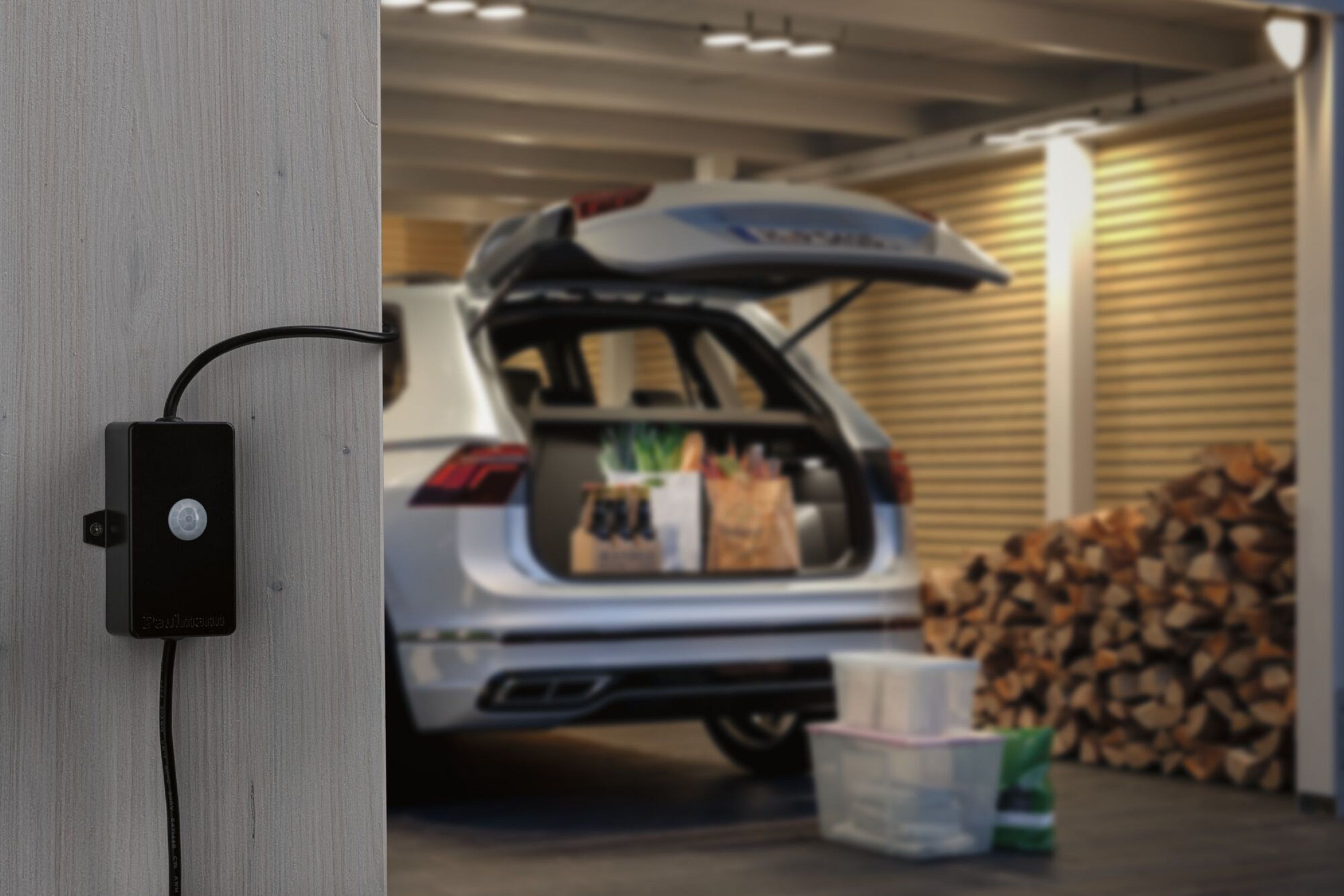 Schwarzer Sensor an Holzwand. Auto mit offenem Kofferraum im Hintergrund. Brennholz sichtbar.