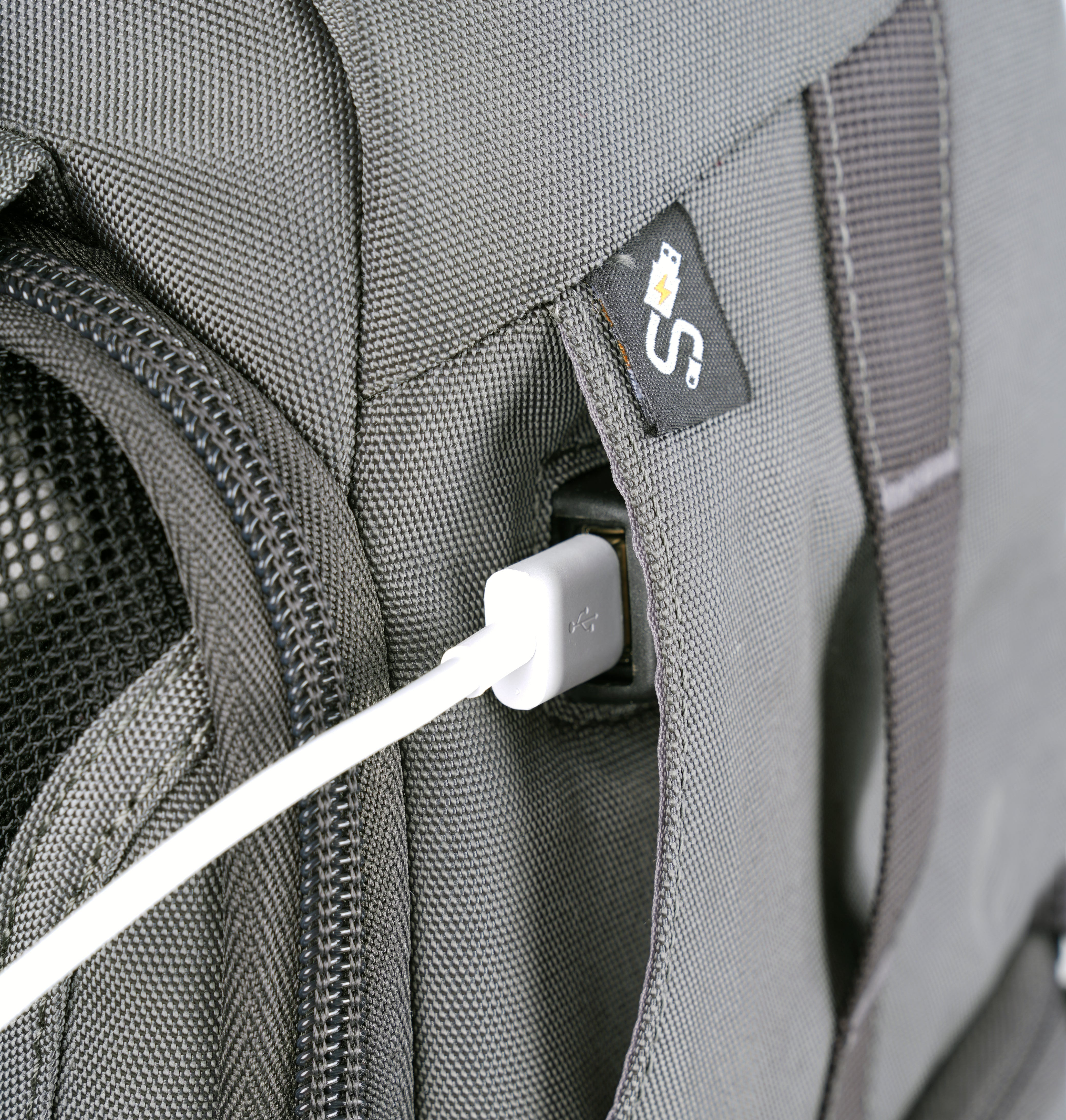 Nahaufnahme eines grauen Rucksacks mit einem weißen USB-Kabel. Ein schwarzer Reißverschluss und ein Logo sind sichtbar.