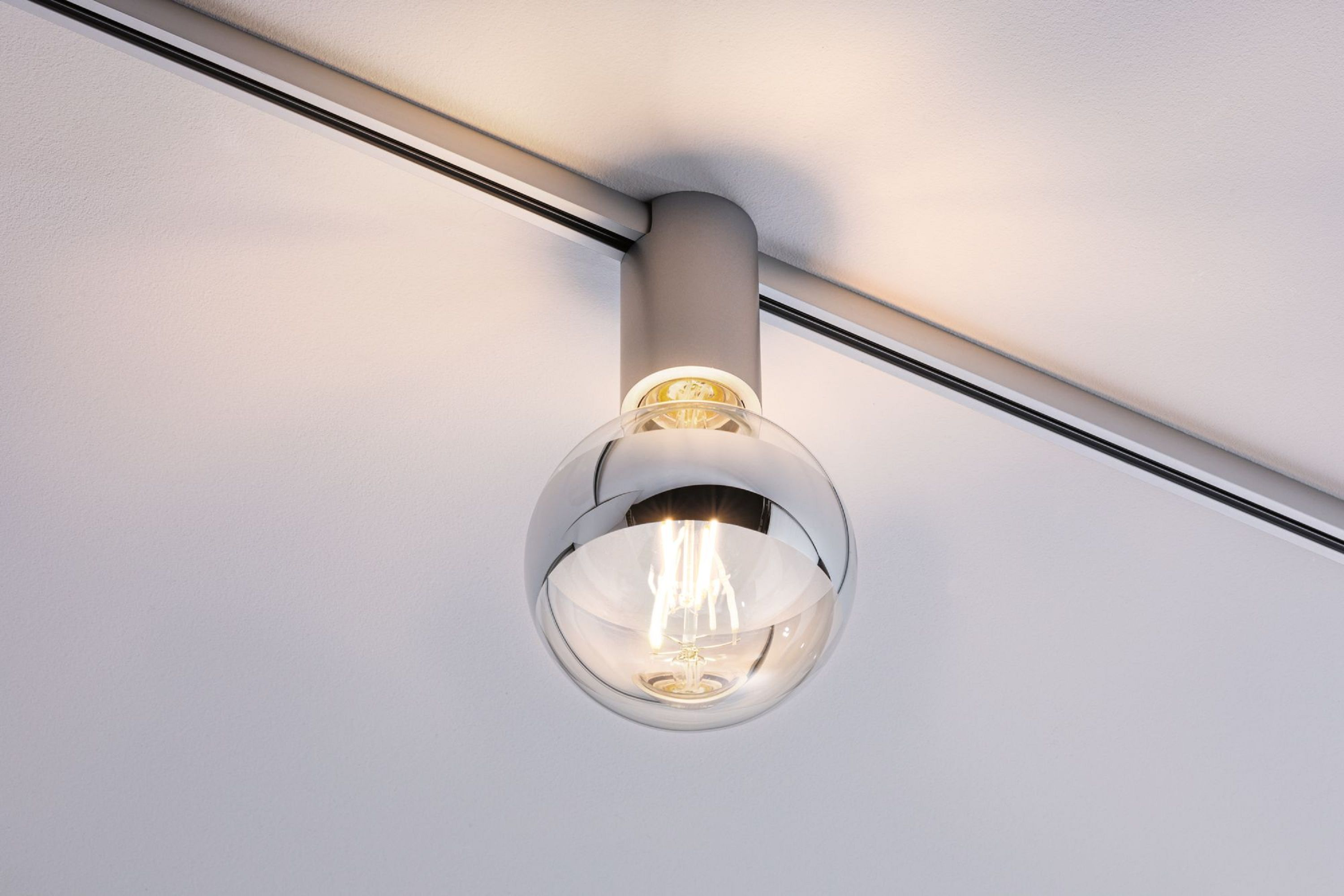 Eine moderne Deckenleuchte mit Glaskugel, silbernem Metallkörper und Schienensystem an einer weißen Decke.