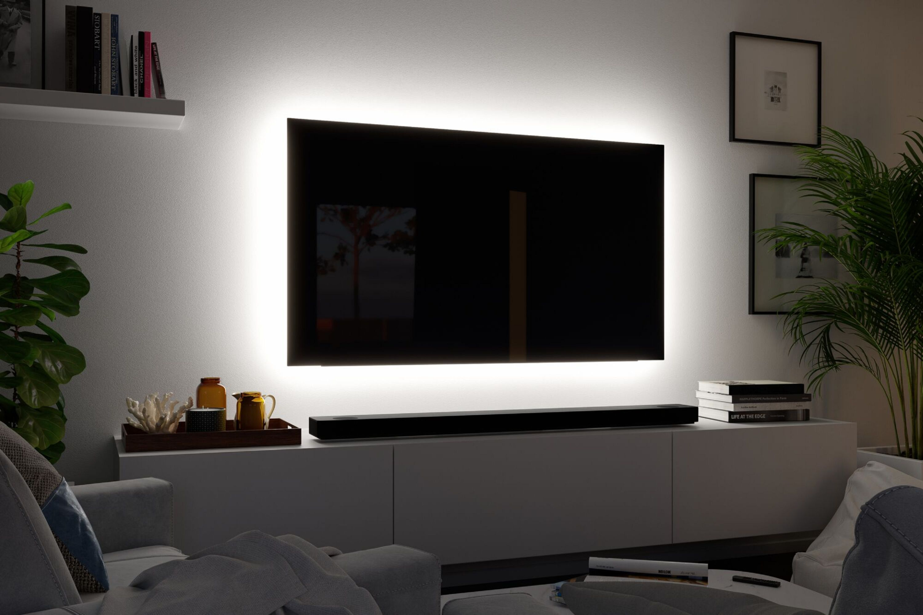 Ein modernes Wohnzimmer mit Fernseher und Soundbar über einem weißen Schrank. Pflanzen und Deko sind sichtbar.