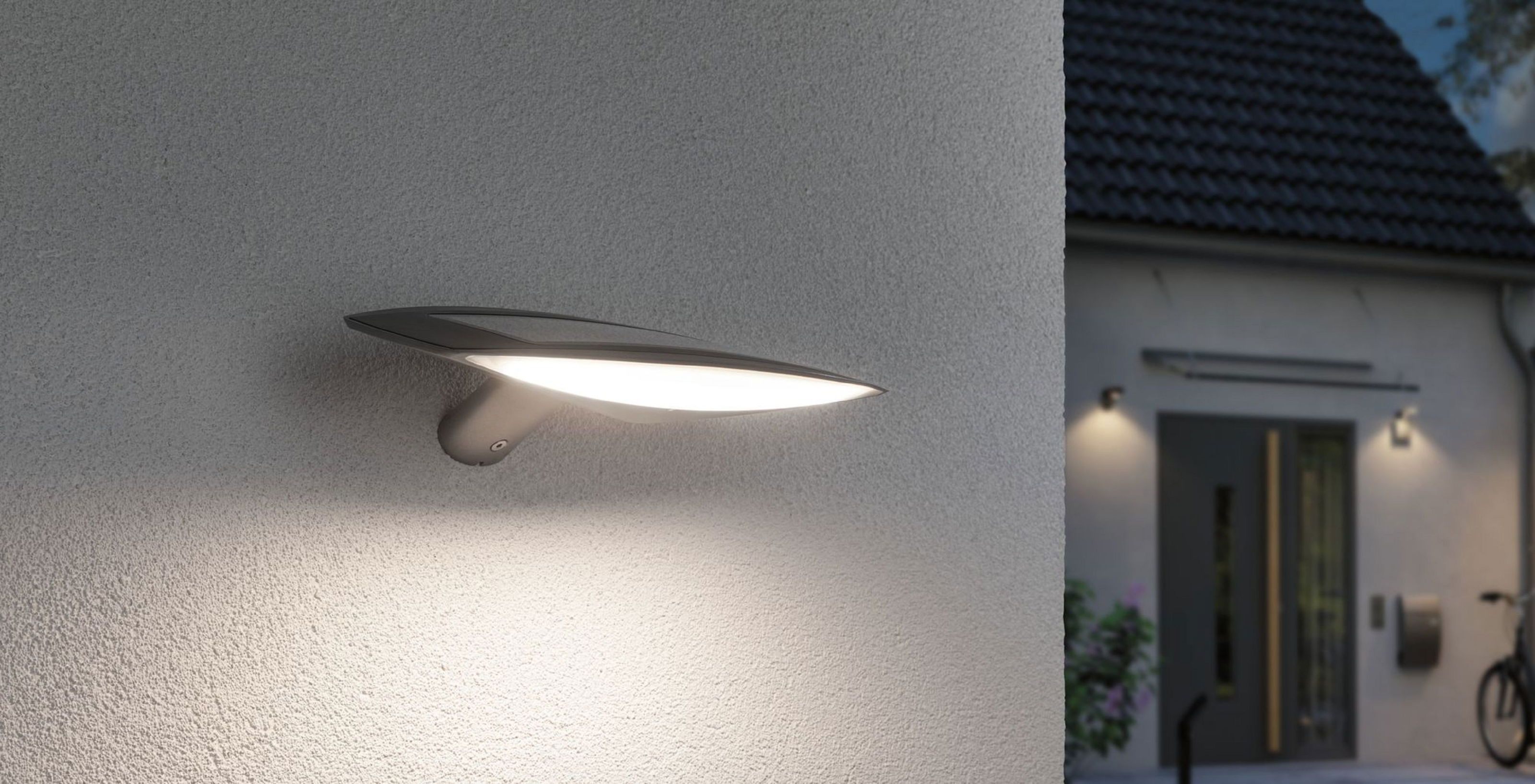 Eine moderne Wandleuchte ist an einer strukturierten grauen Wand montiert. Im Hintergrund befindet sich ein Haus. Die Lampe beleuchtet die Wand.