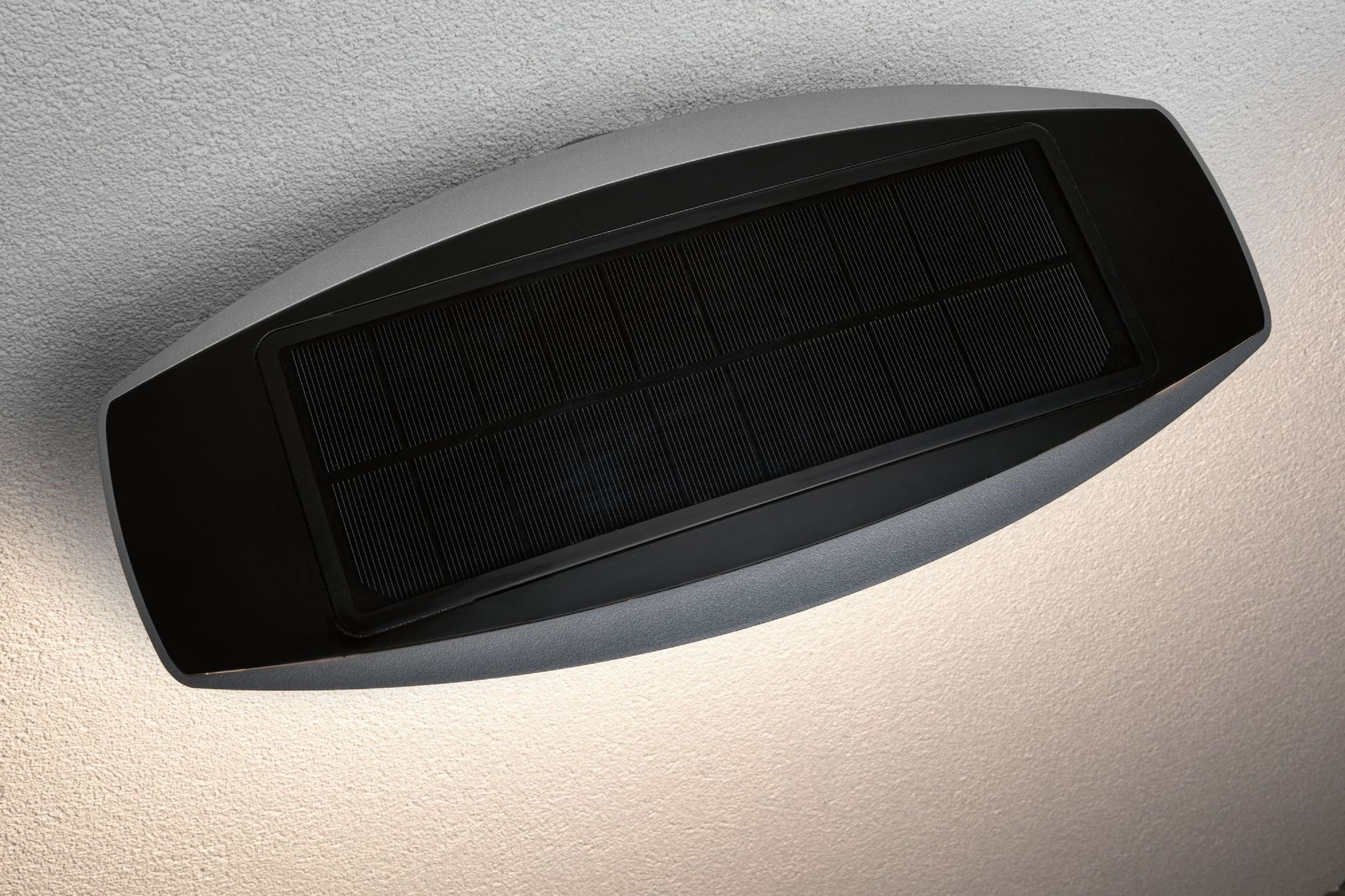 Ein schwarzes Solarpanel befindet sich auf einer grauen Leuchte, die an einer strukturierten, hellen Wand montiert ist.