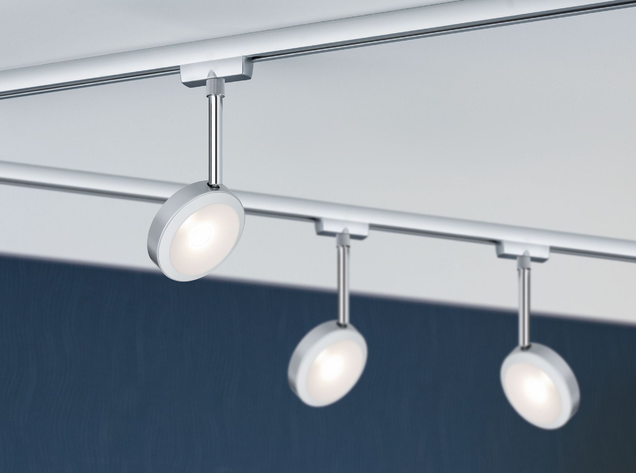 Drei moderne Deckenstrahler. Sie sind silber und weiß und befinden sich auf einer weißen Schiene vor blauem Hintergrund.