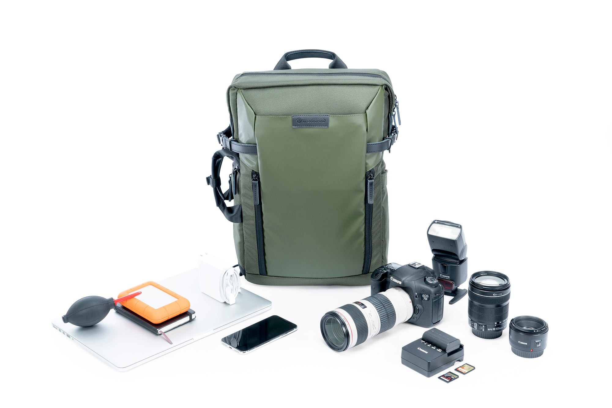 Ein olivgrüner Kamerarucksack, Laptop, Telefon, Kamera, Objektive, Blitz und Zubehör sind auf einer weißen Oberfläche angeordnet.